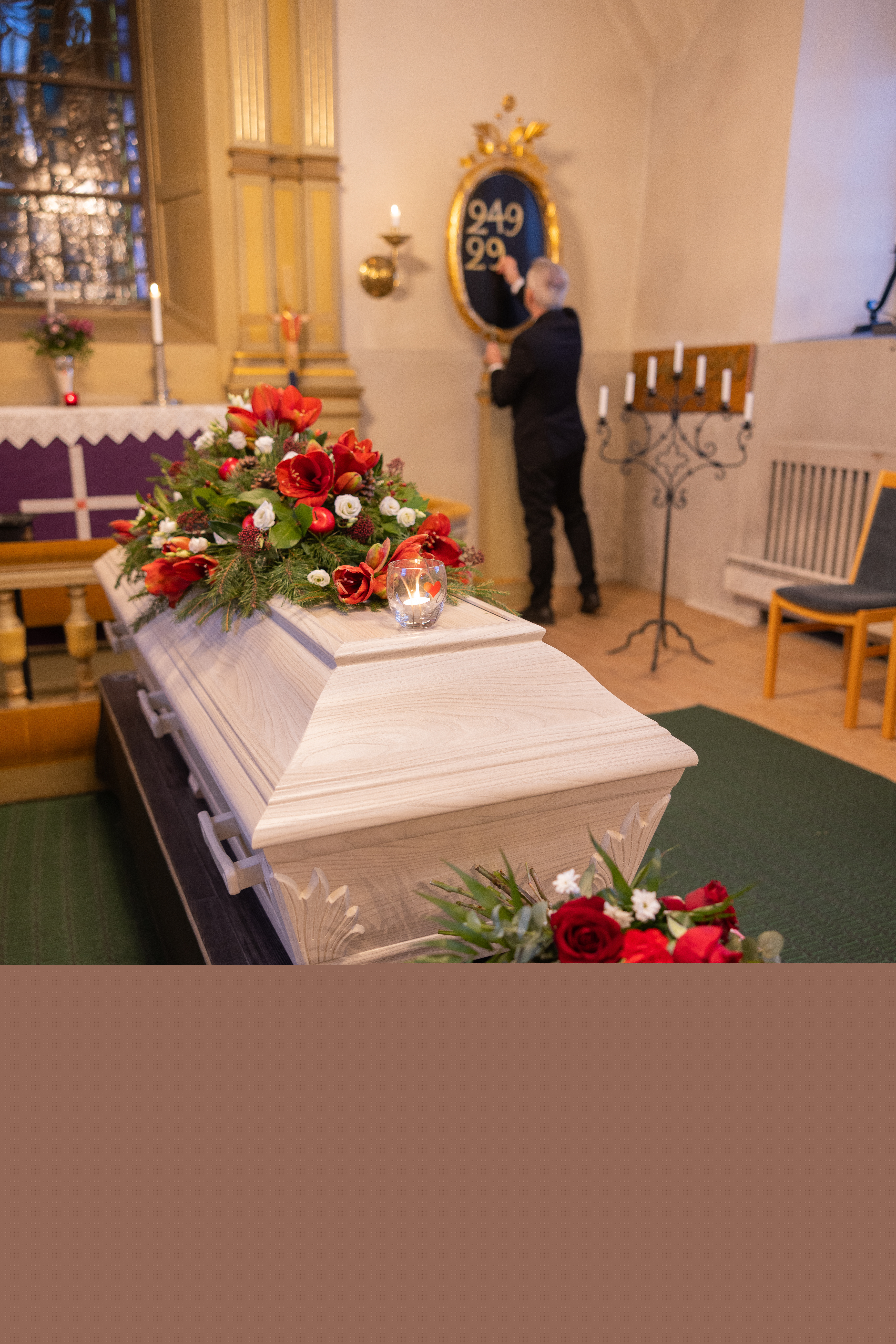 En kyrkvaktmästare hänger upp siffror på psalmtavlan. I koret står en vit kista med röda och vita blommor på.