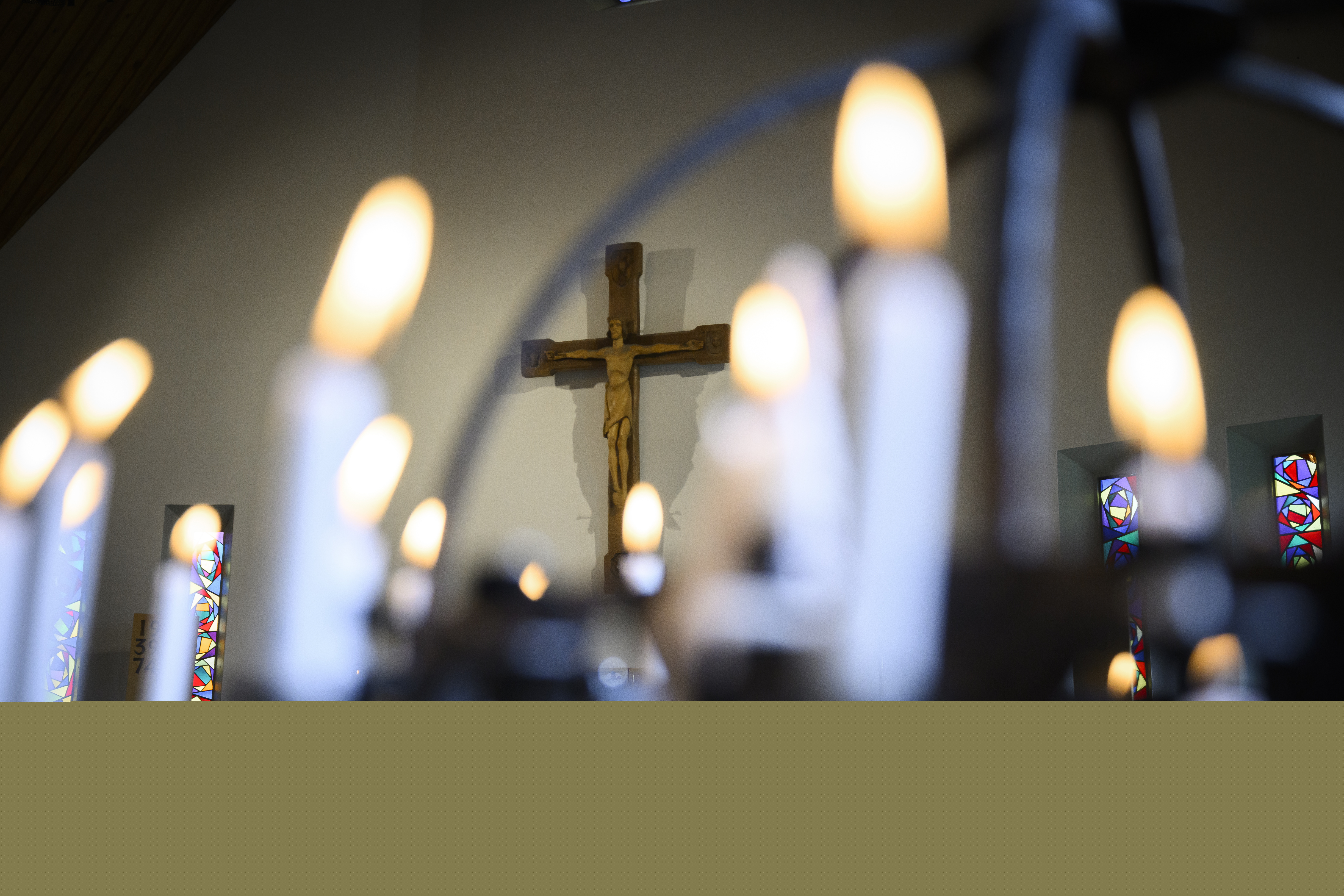 Ett krucifix är upphängt på en vit vägg. Flera tända ljus i en ljusbärare i förgrunden.