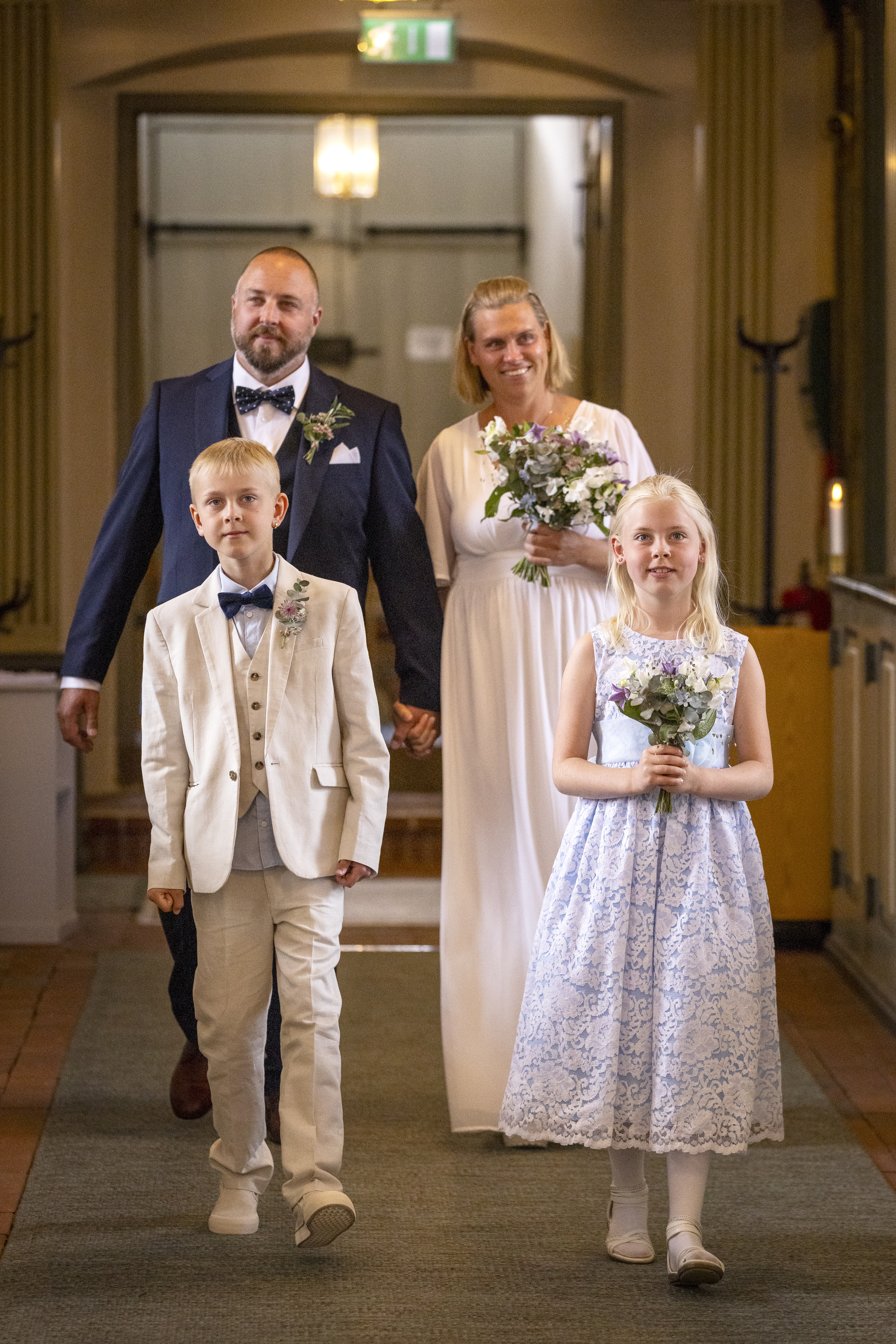 Drop-in vigsel i en kyrka. Ett brudpar och två yngre barn går i altargången. Bruden och ett av barnen håller i varsin blombukett.