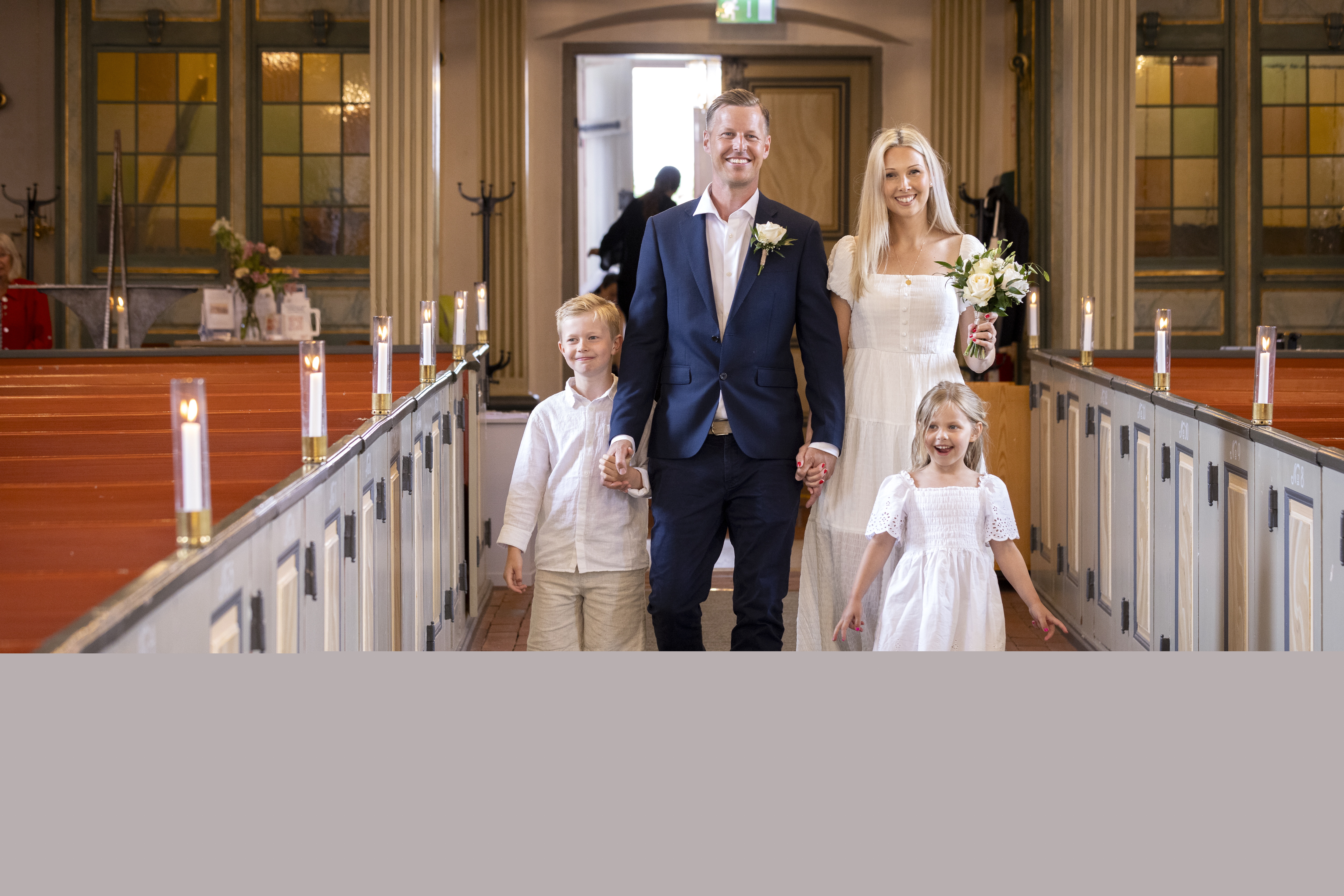 Drop-in vigsel i en kyrka. Ett brudpar går i altargången tillsammans med två ljusklädda barn. Bruden håller en vit blombukett i handen.