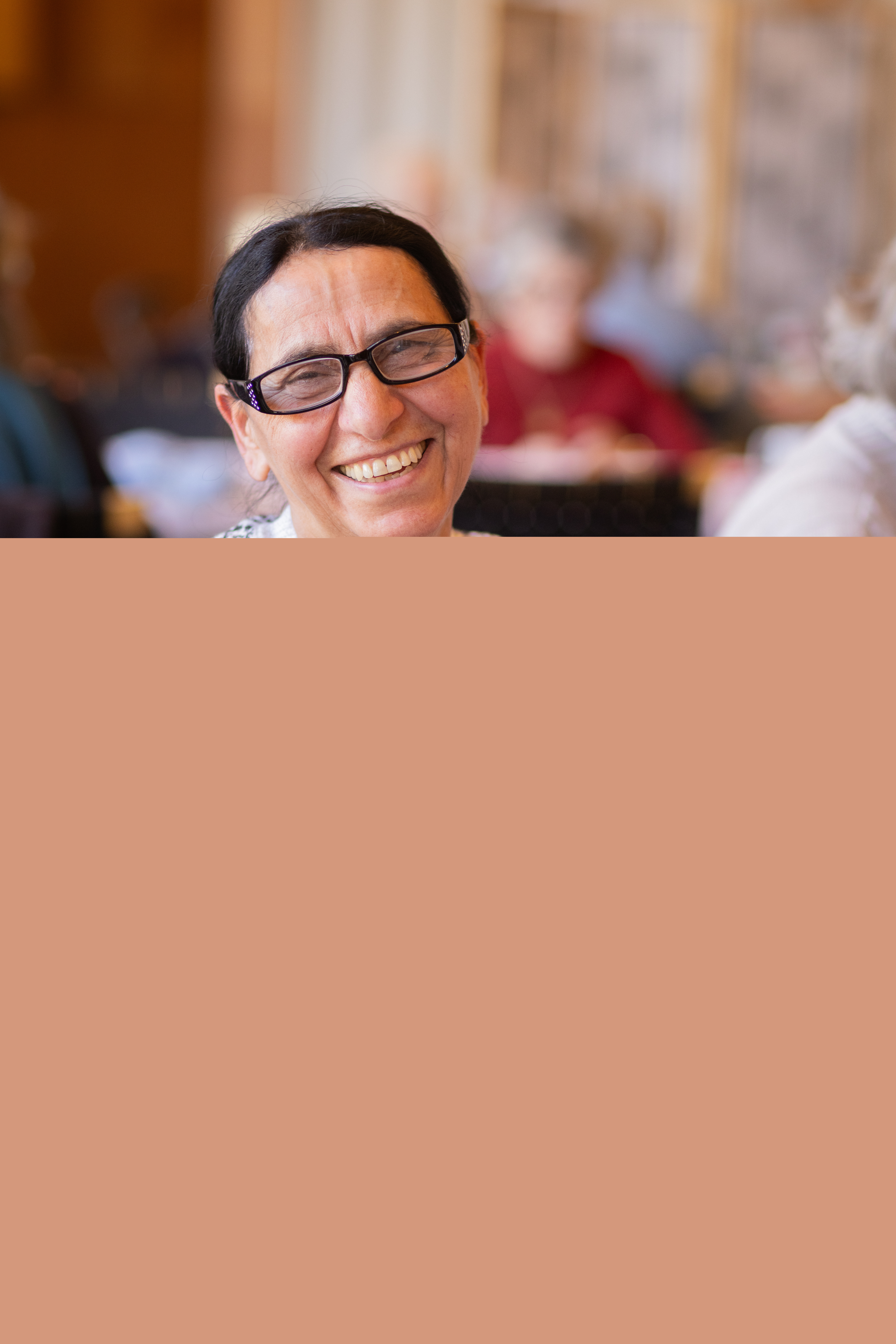 En kvinna sitter på en stol och stickar. Hon ler mot kameran.