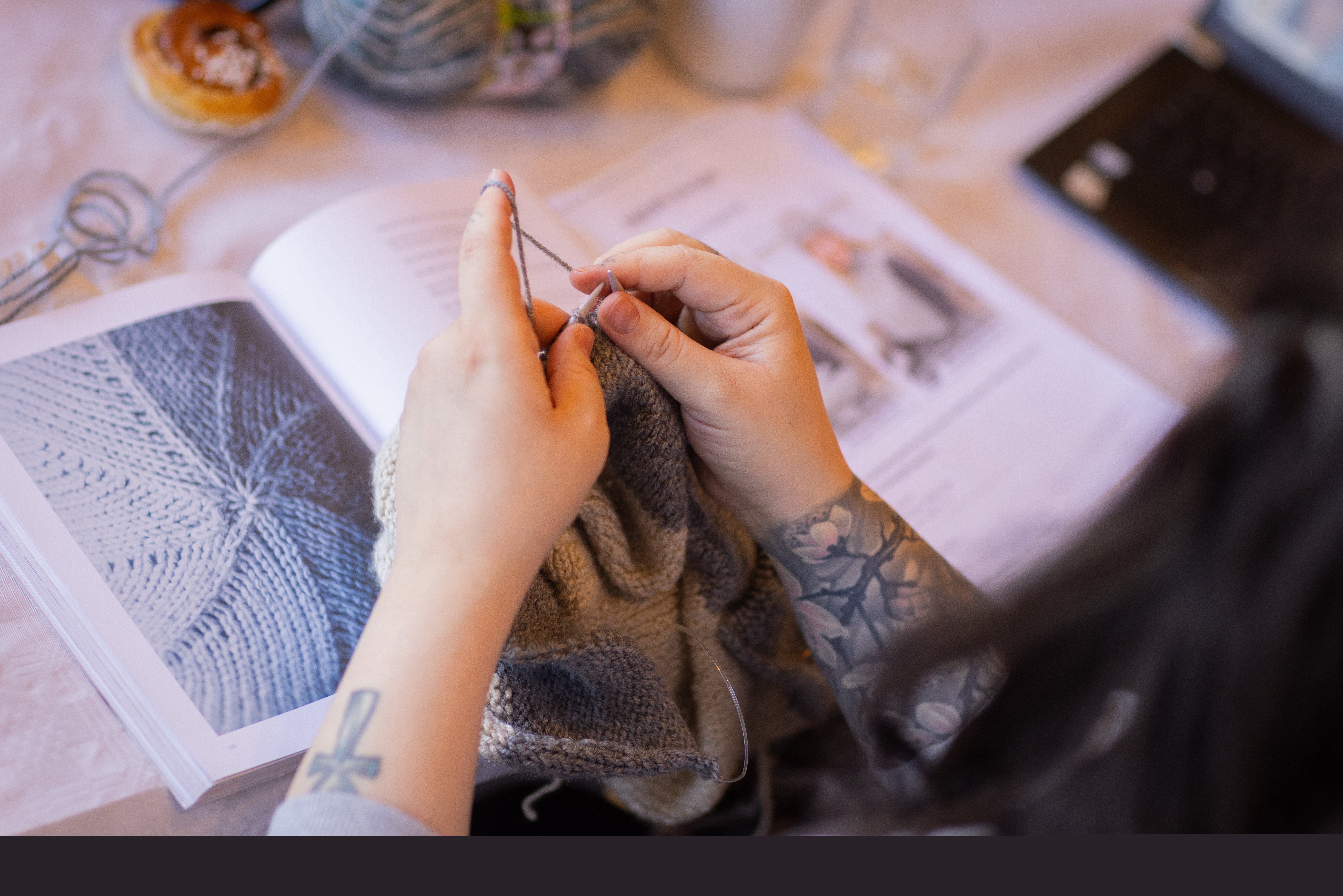 En person sitter vid ett bord och stickar. På bordet ligger en instruktionsbok i stickning.