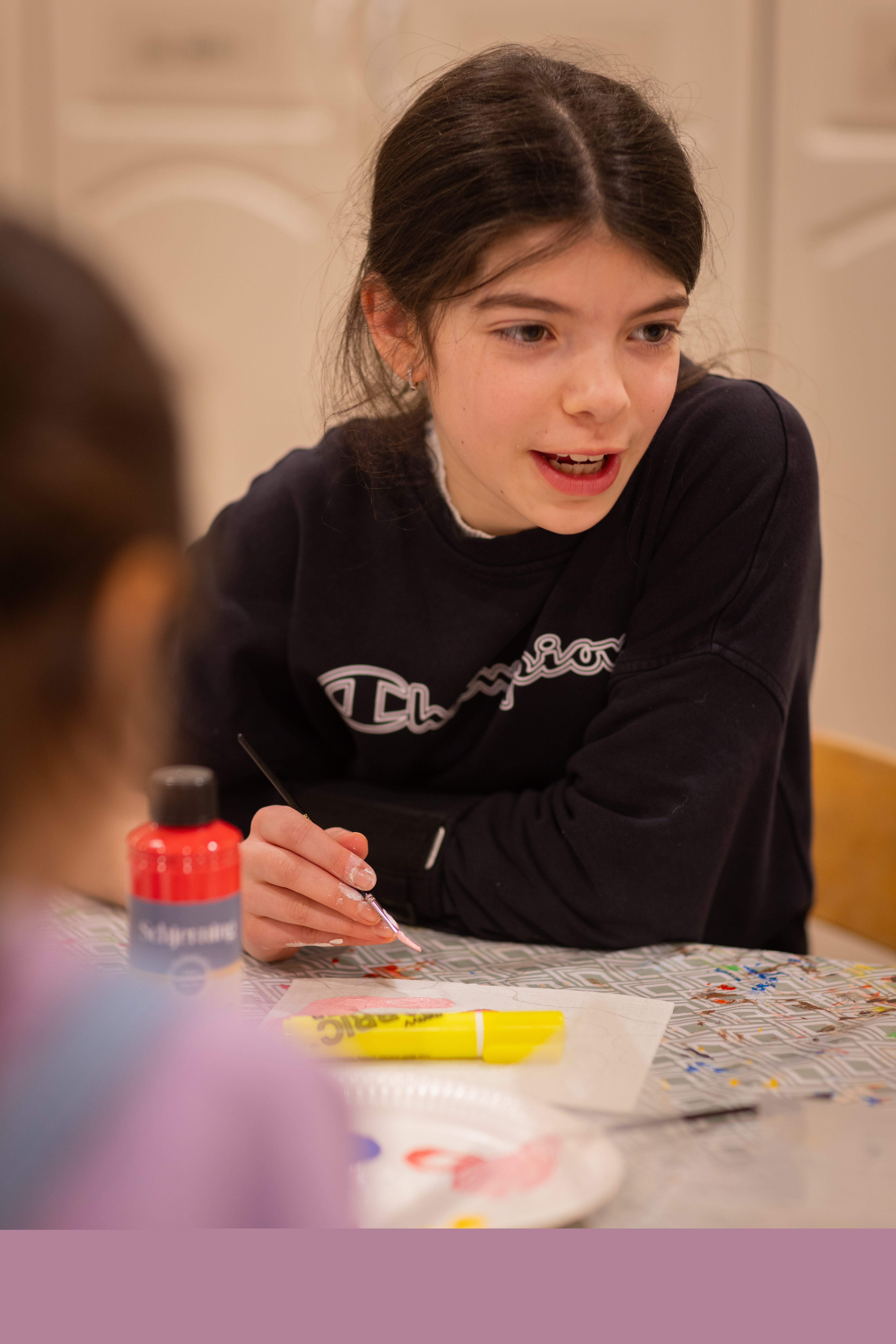 En flicka sitter vid ett bord och målar med pensel. Ett annat barn sitter i förgrunden.