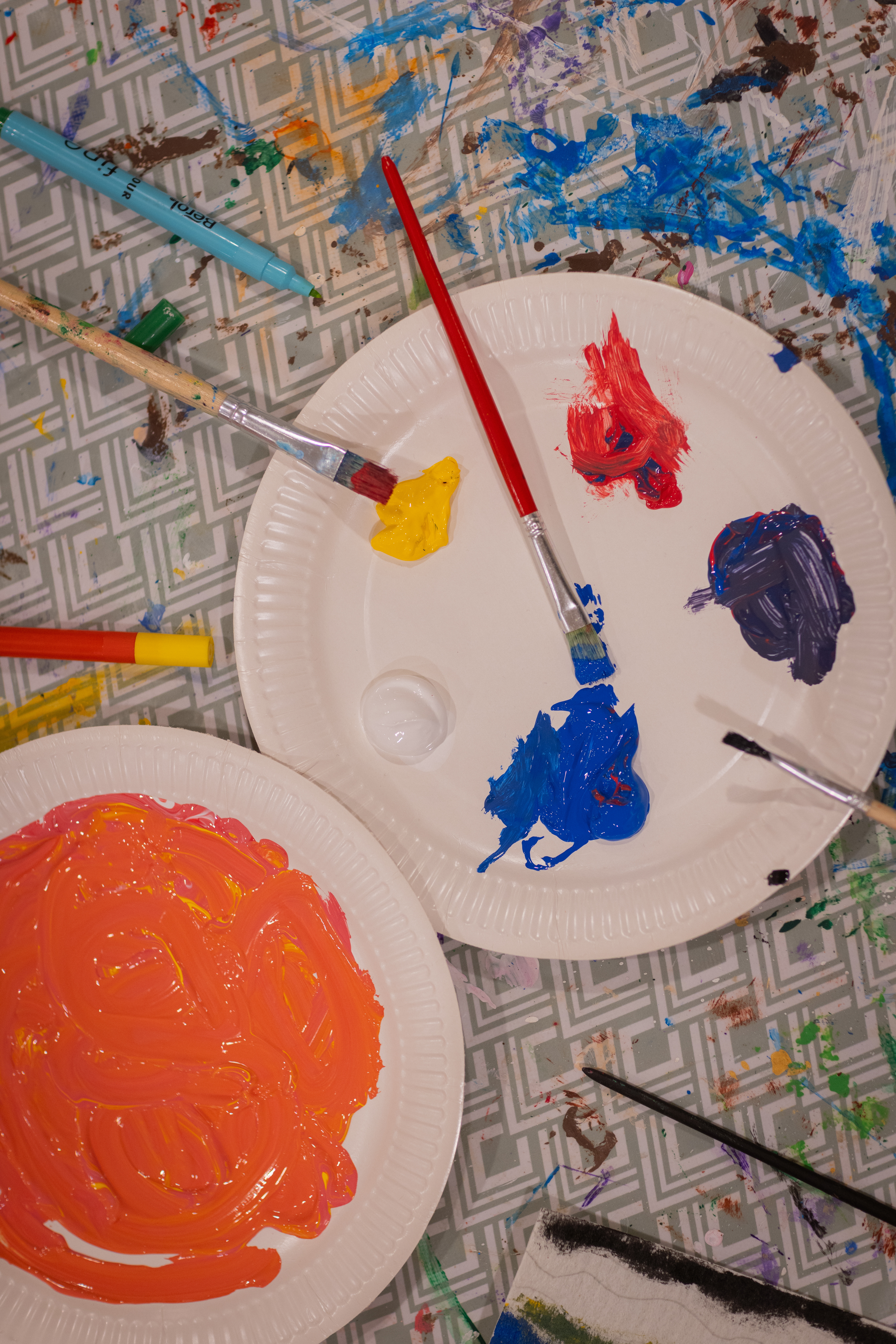 Två papperstallrikar med målarfärg på står på ett bord. Målarpenslar i olika storlekar ligger i närheten.