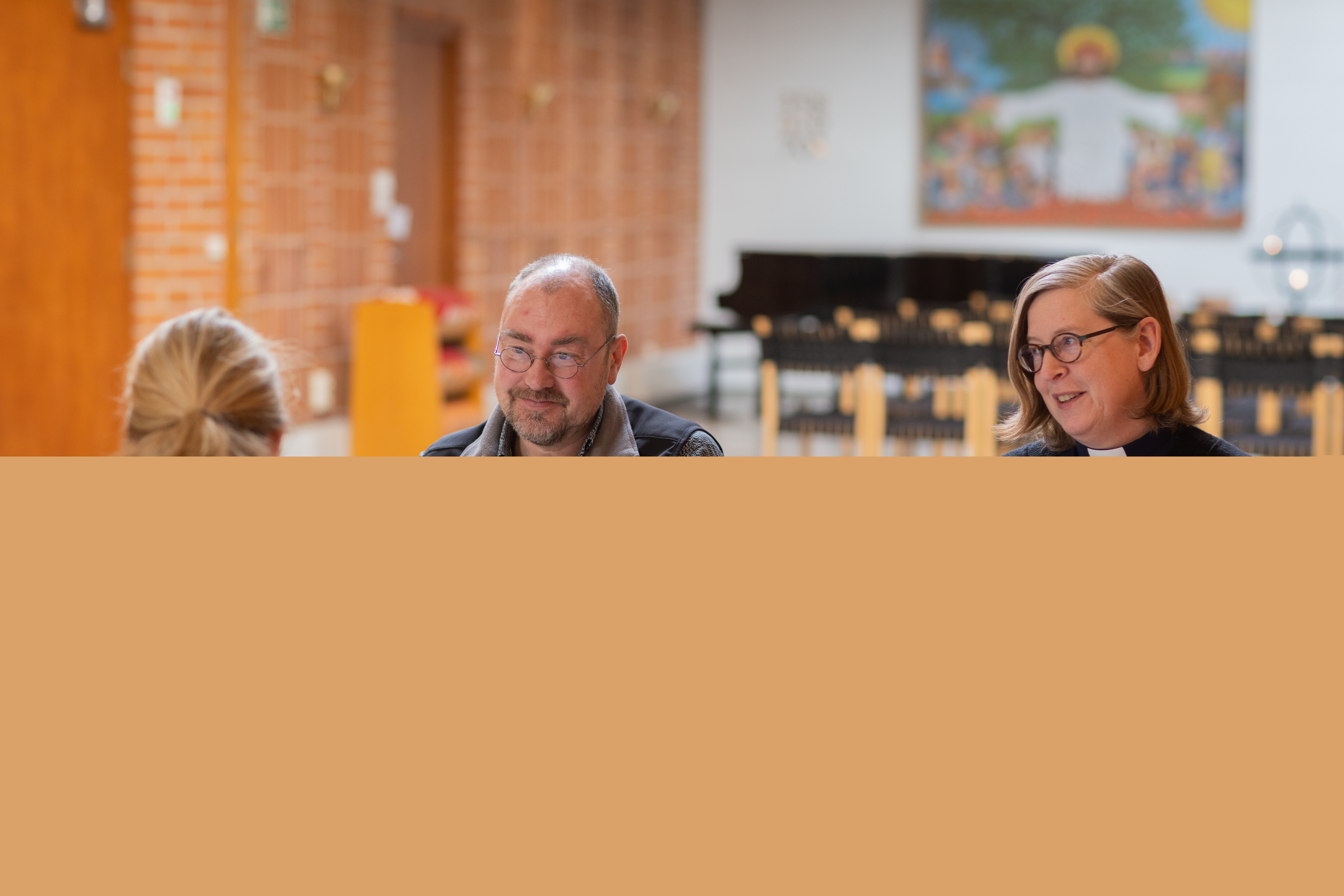 En präst sitter vid ett bord och samtalar med två andra personer.