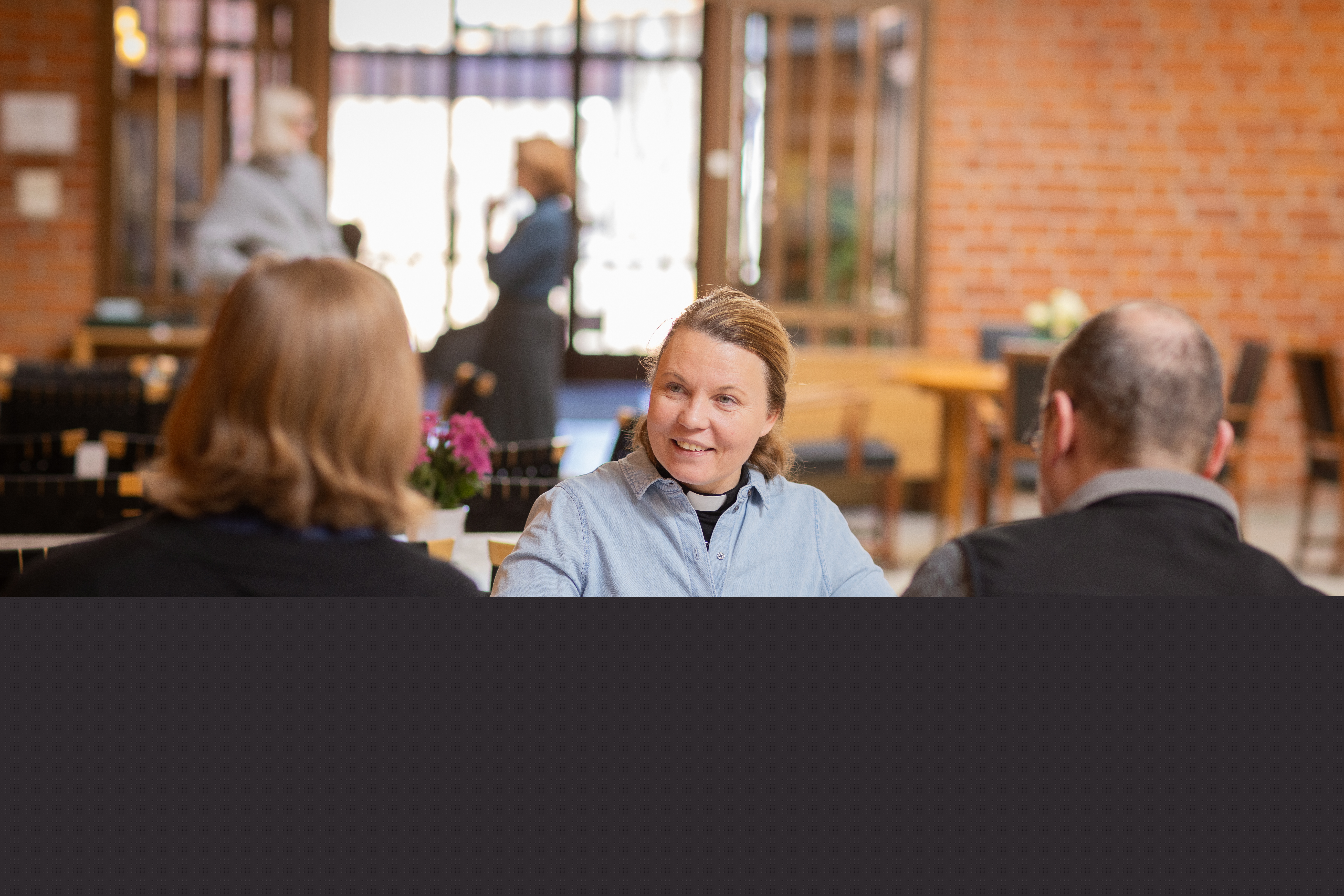 En kvinnlig präst sitter vid ett bord och samtalar med två andra personer.
