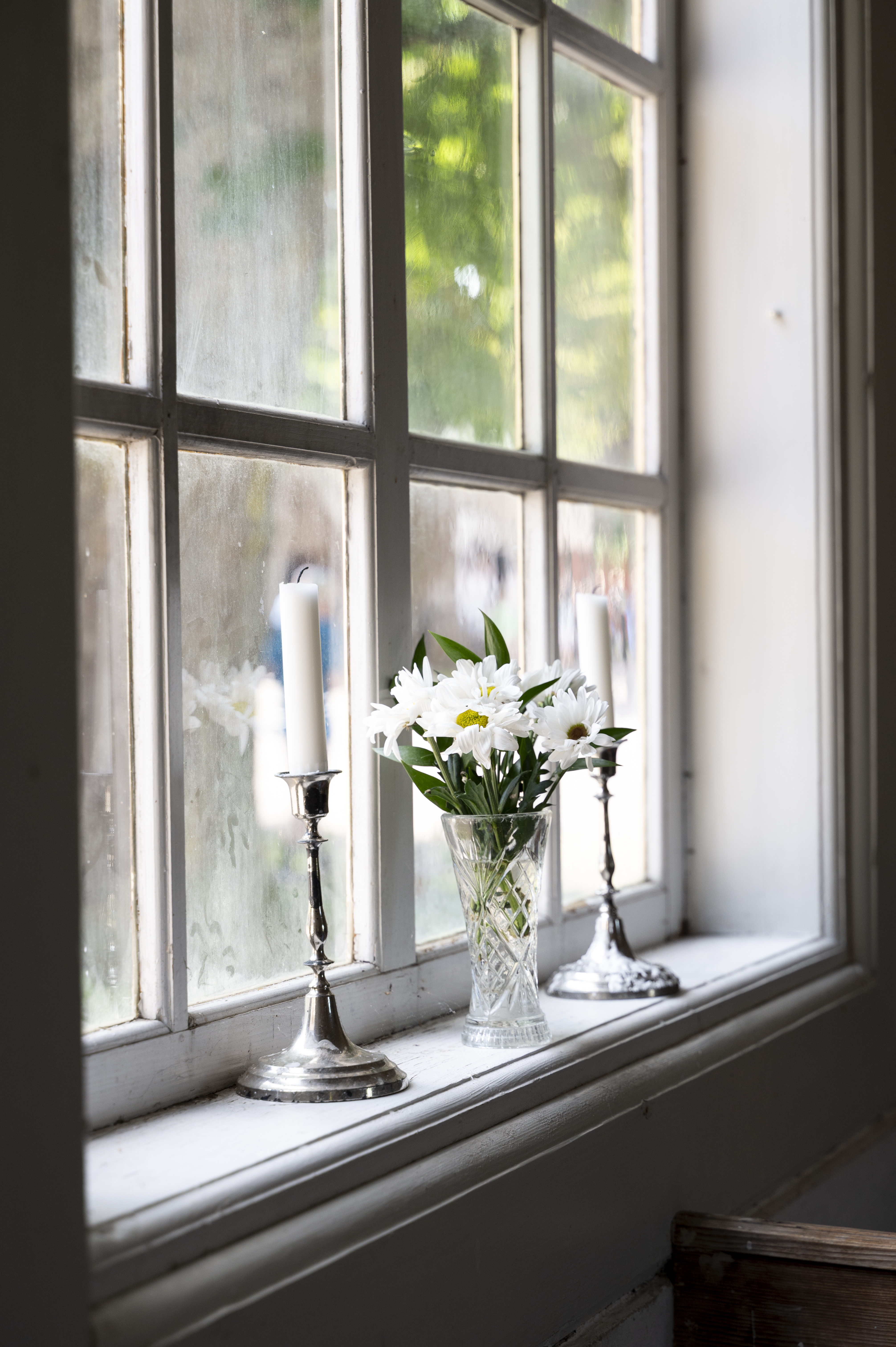 Två silverljusstakar och en vas med vita blommor står i ett fönster.