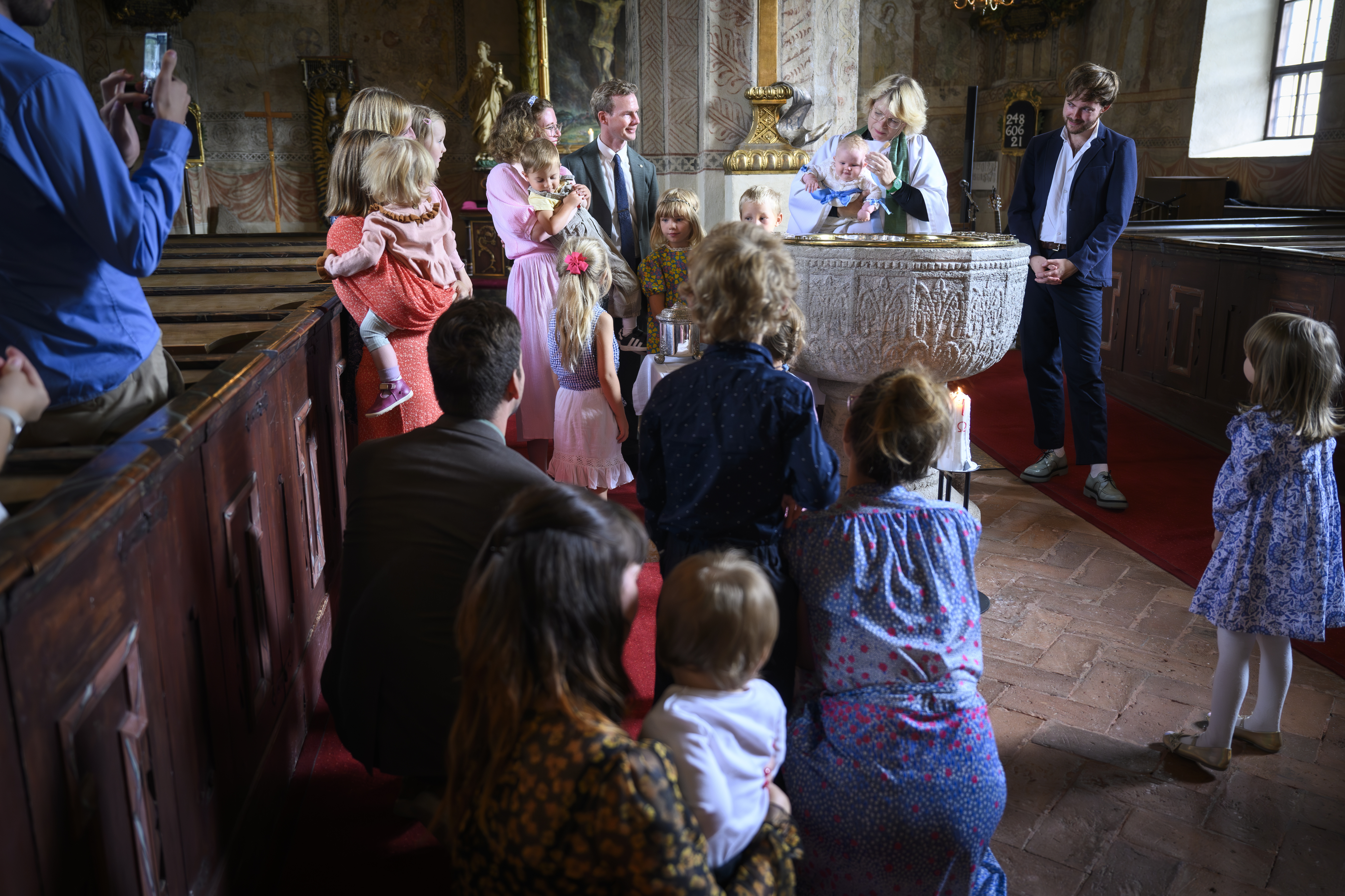 En präst döper ett barn. Flera personer med barn i famnen står vid dopfunten.
