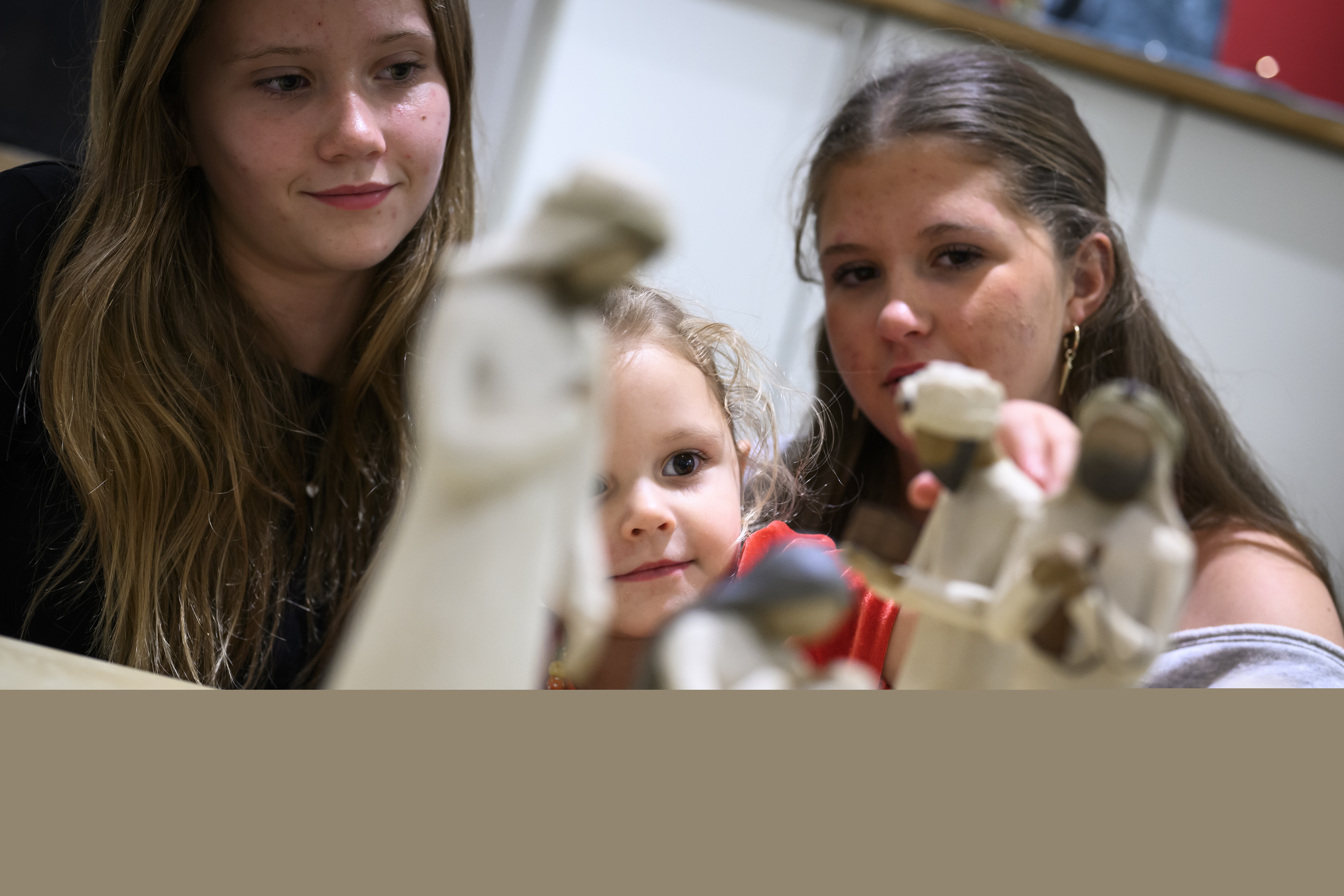 Trädockor som föreställer Jungfru Maria med Jesus-barnet i famnen och de tre vise männen står på ett bord. Ett barn och två ungdomar sitter runt bordet.