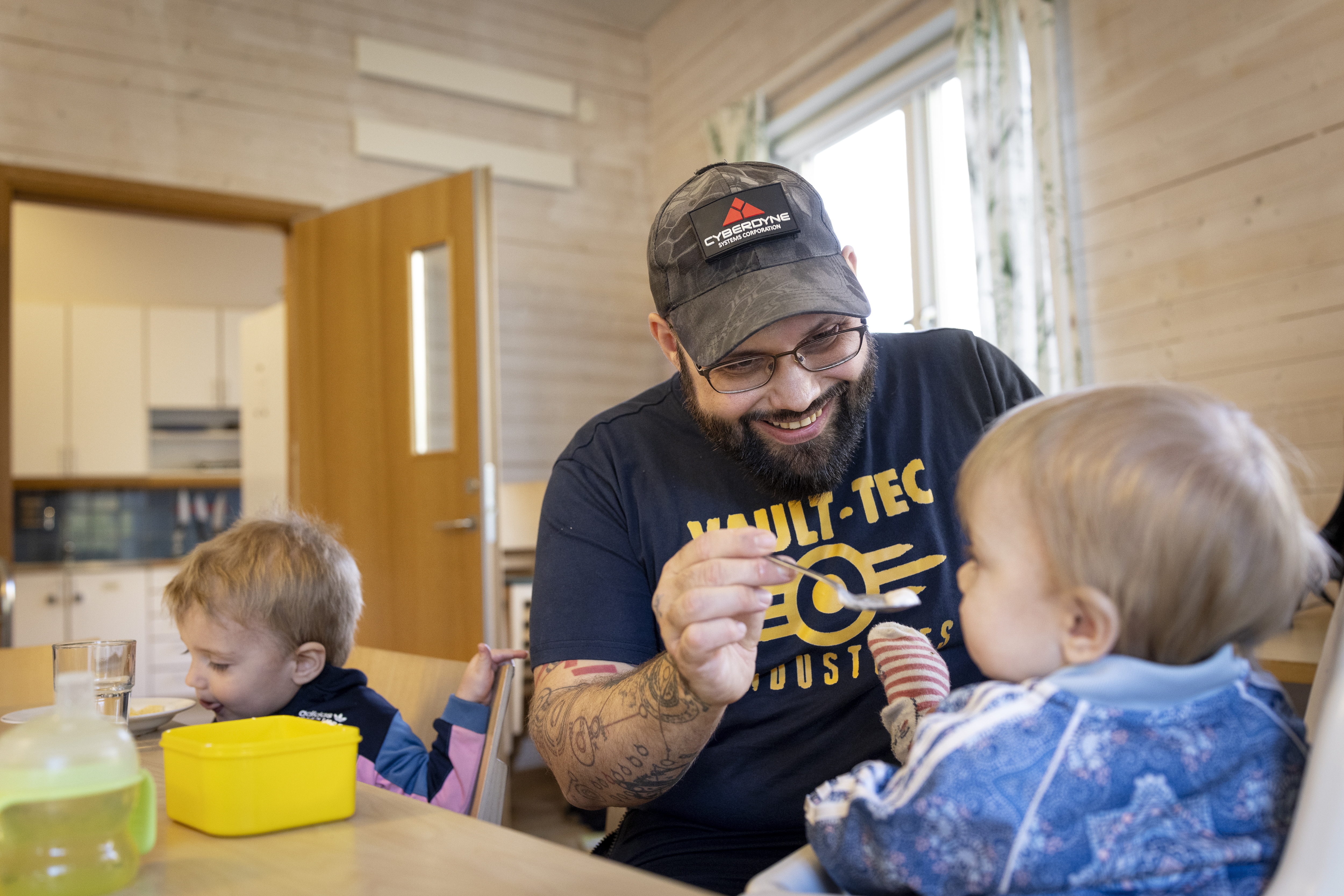 En leende man sitter vid ett bord och matar ett litet barn med sked. Ett annat barn sitter bredvid och äter.