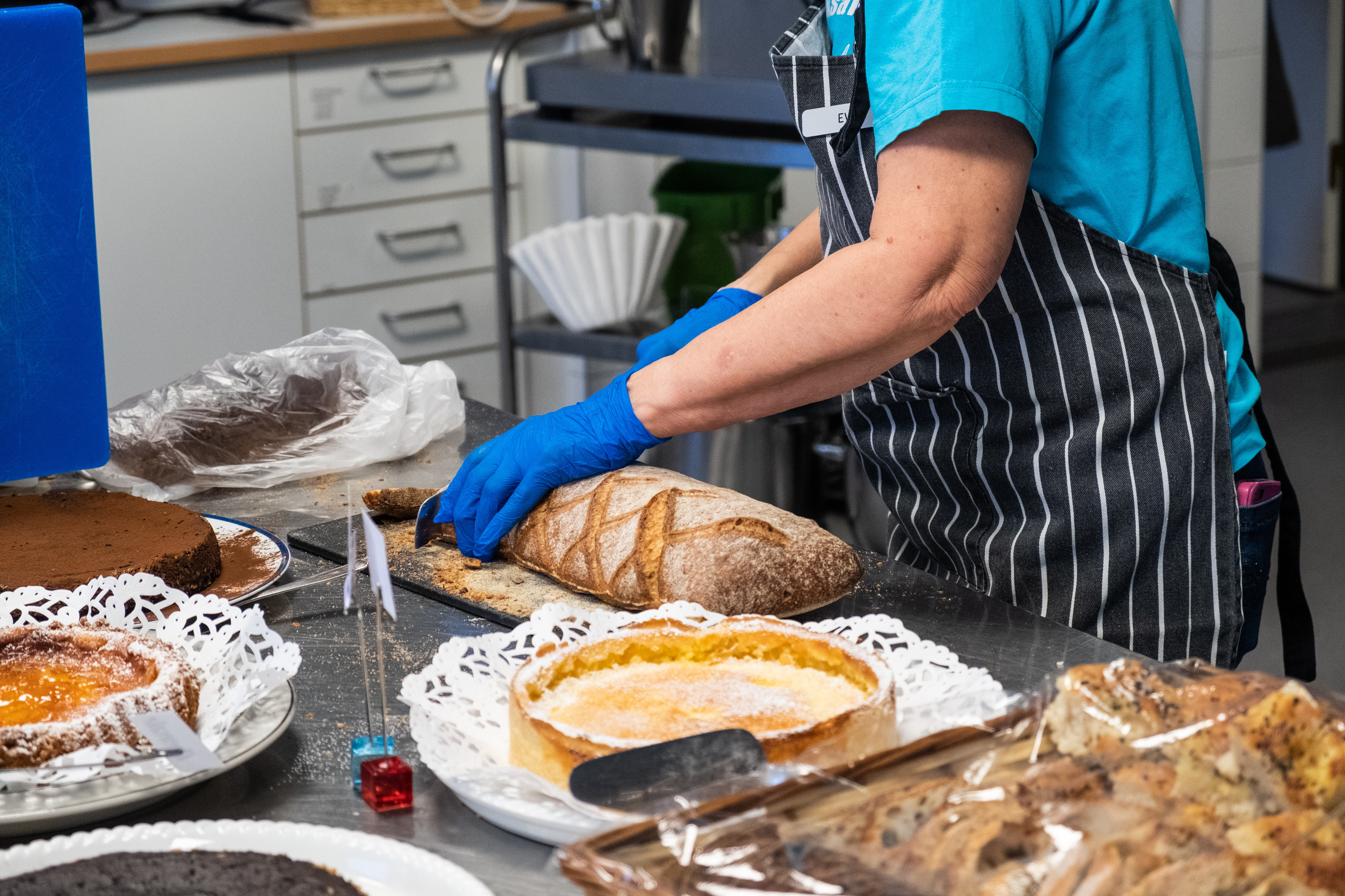 En person med förkläde och plasthandskar står vid ett bord med bakverk och skär upp bröd.