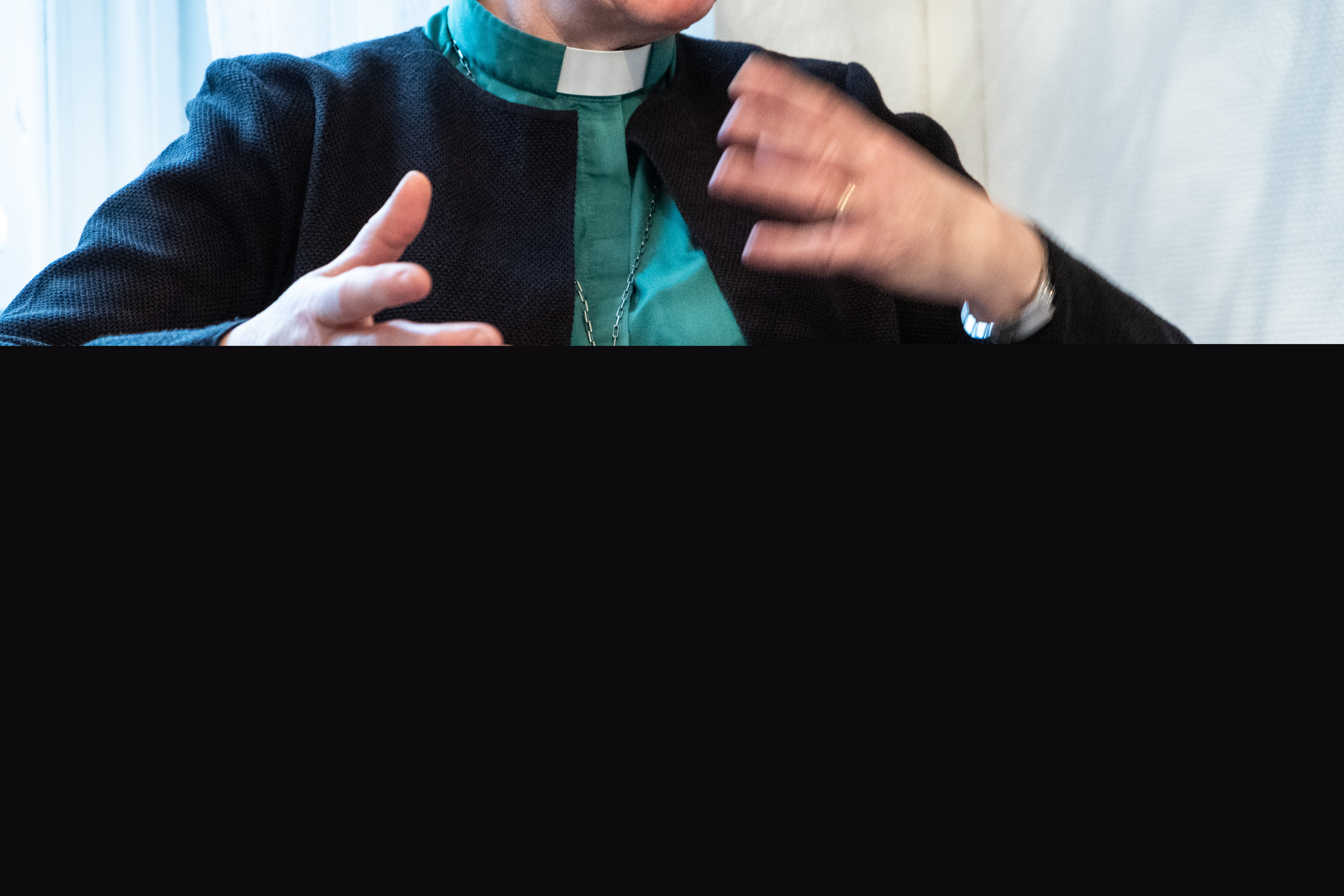 En diakon sitter i en fåtölj och gestikulerar. Hen bär diakonemblemet runt halsen.