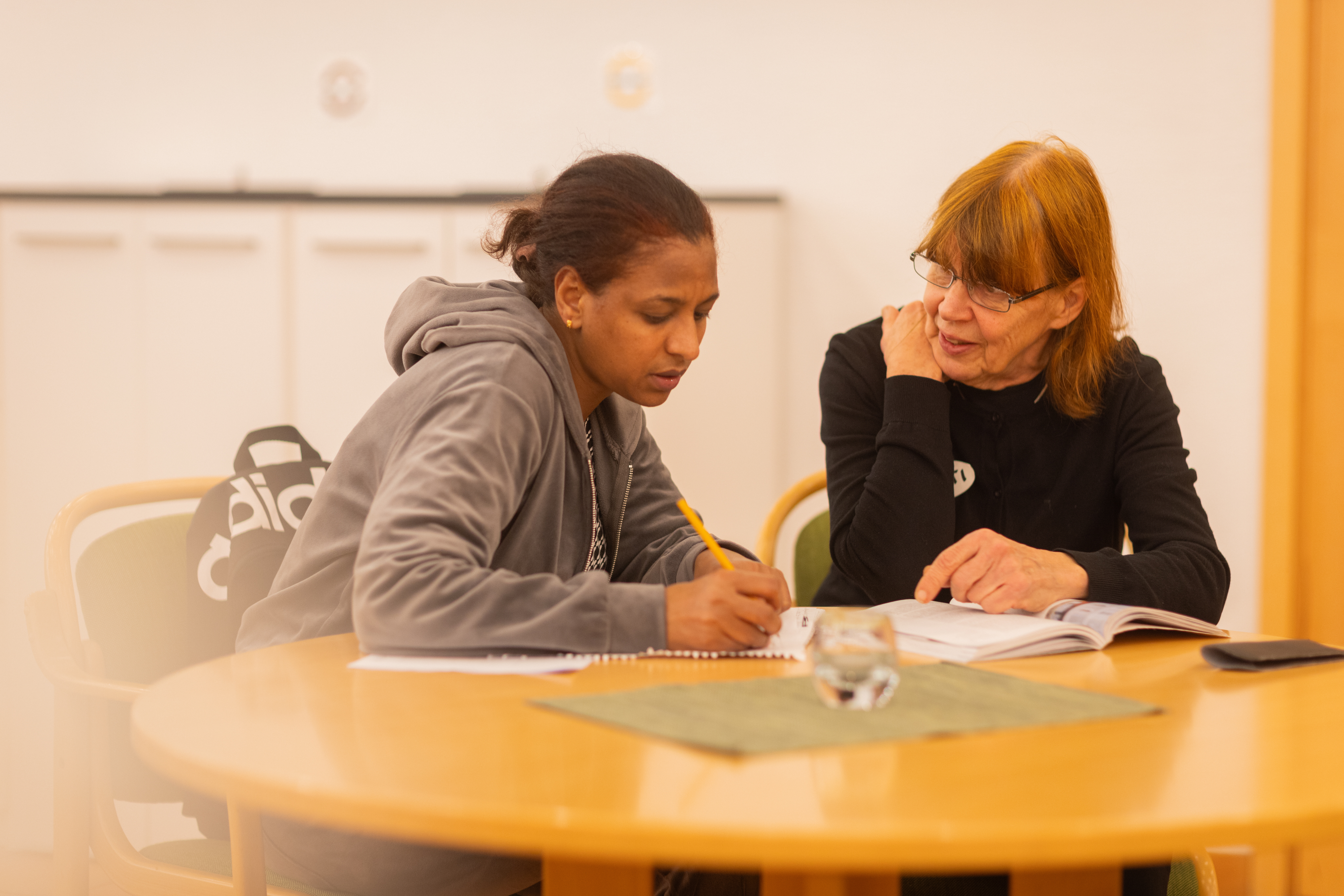 Två kvinnor sitter vid ett bord. En av kvinnorna håller en penna i handen, den andra har en bok framför sig.