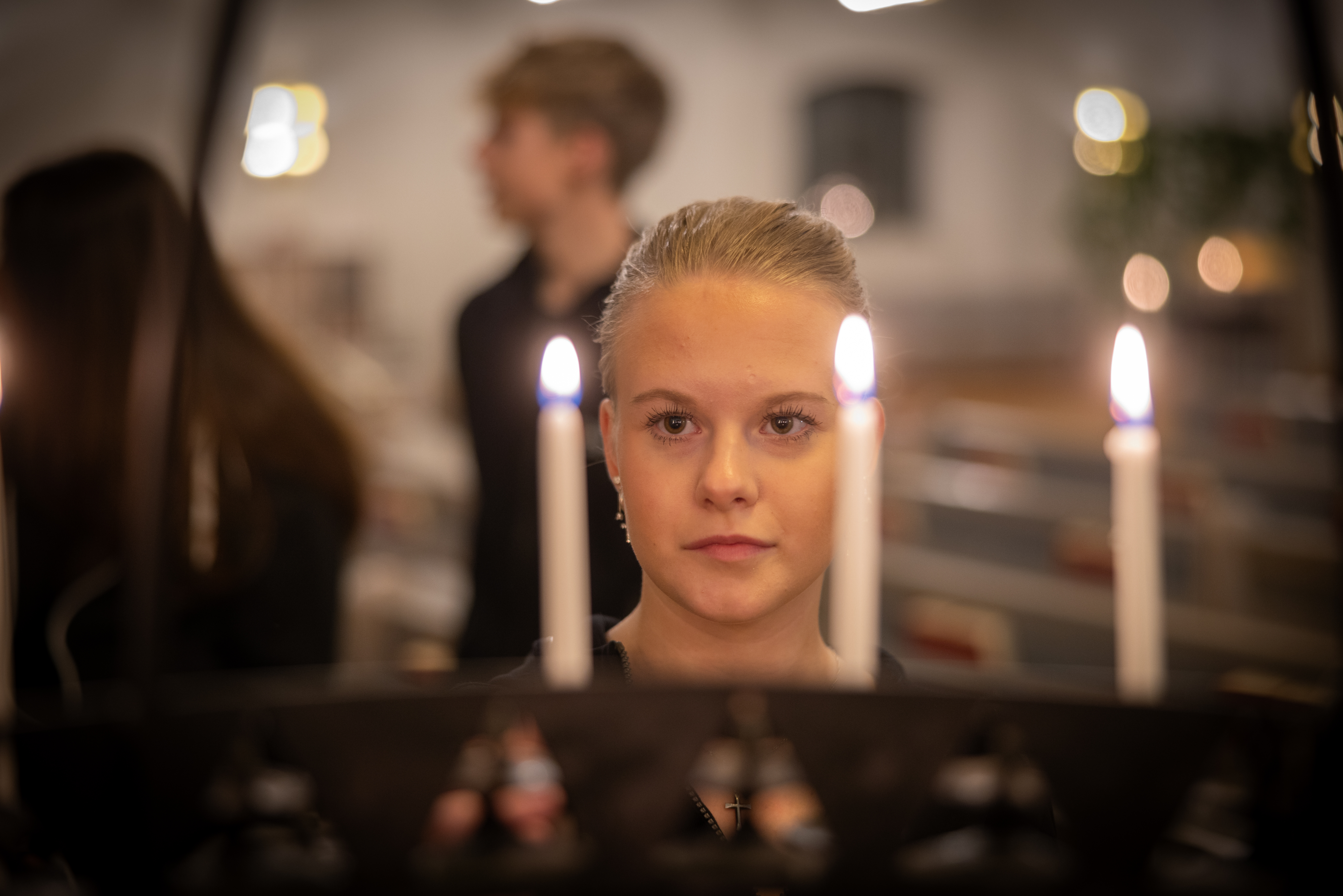 En tjej står framför en ljusbärare med tända ljus. I bakgrunden står några ungdomar.