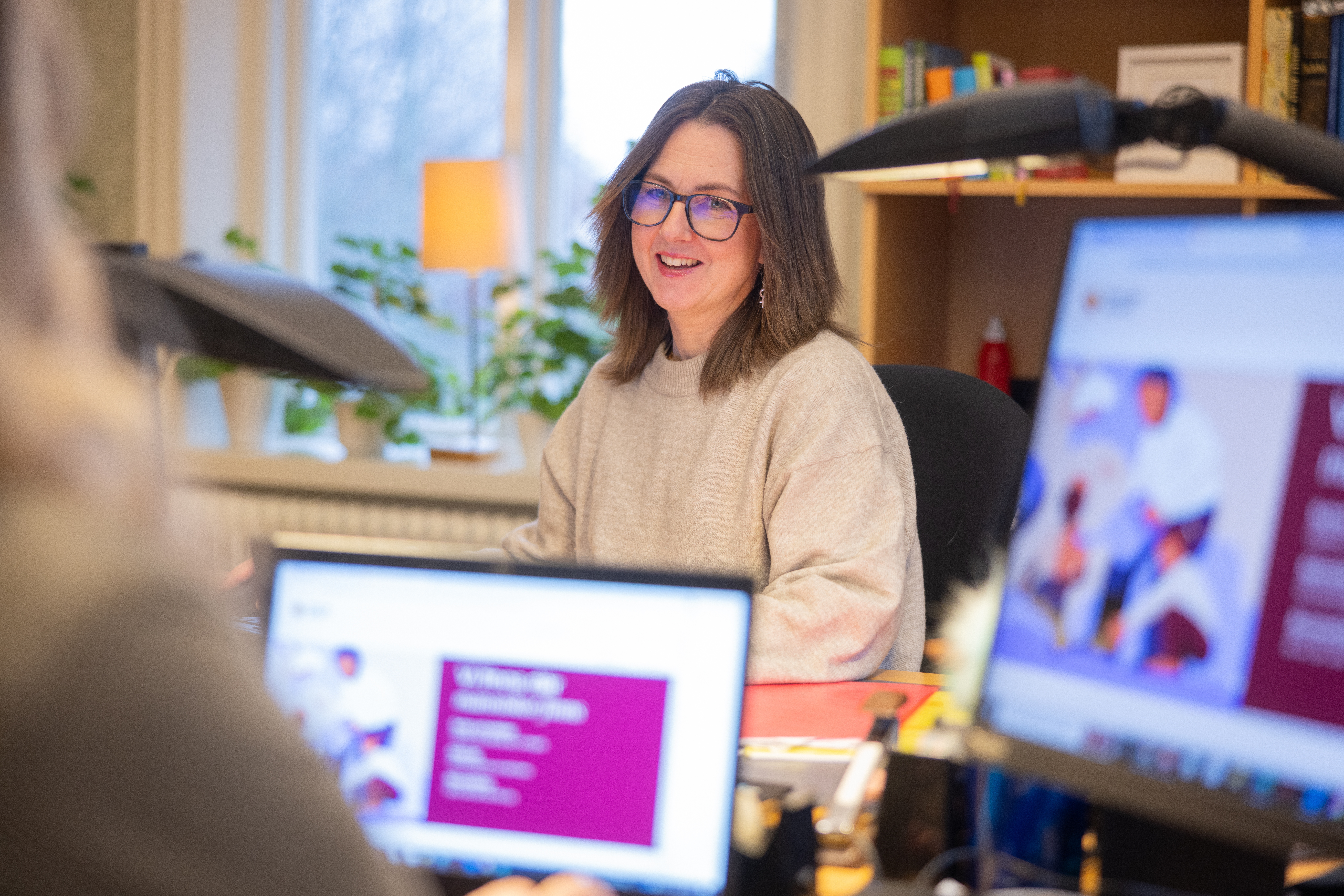 En kvinna i beige tröja sitter och arbetar på ett kontor. Hon pratar med en kollega.