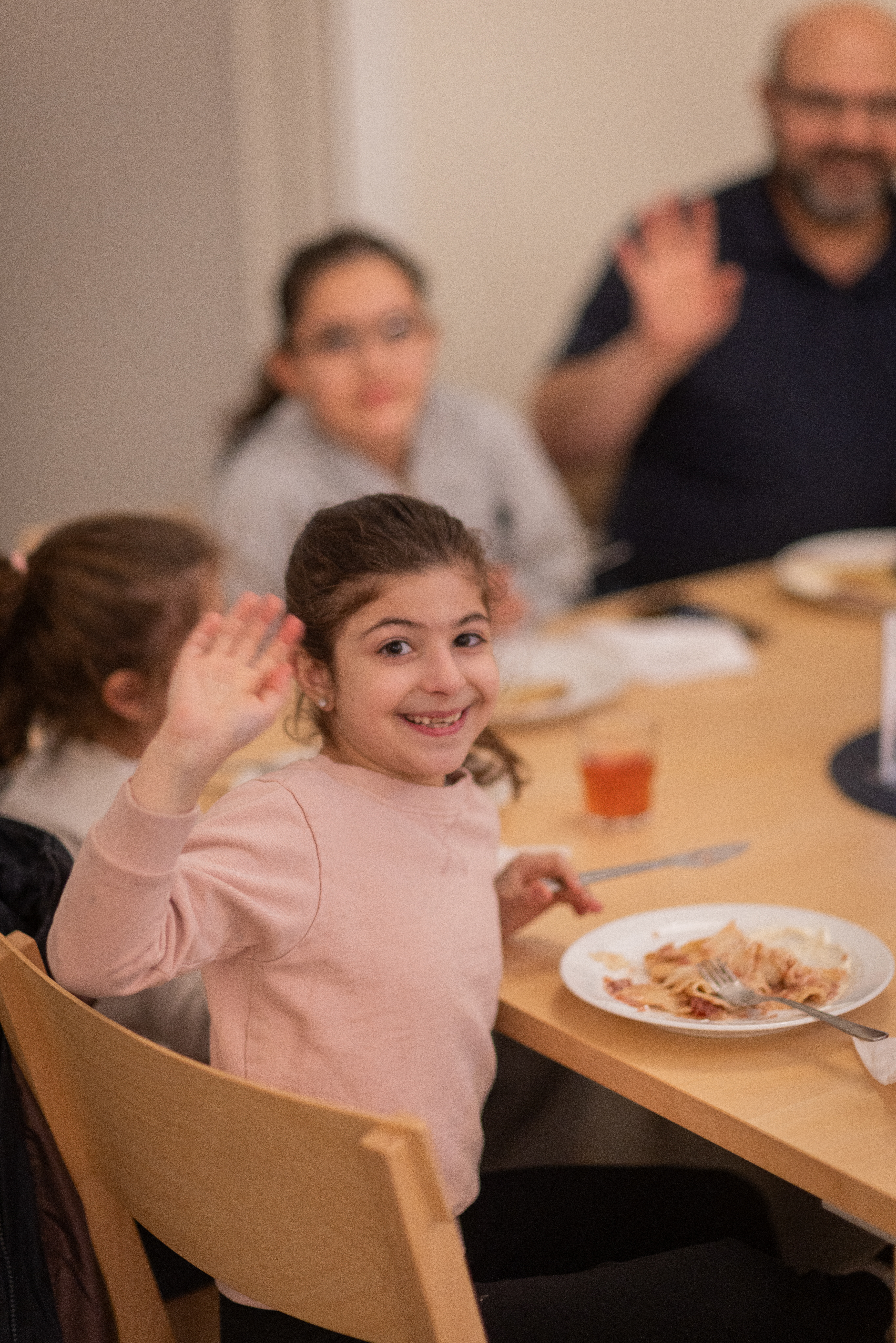 En flicka som sitter och äter vid ett bord vänder sig mot kameran och vinkar.