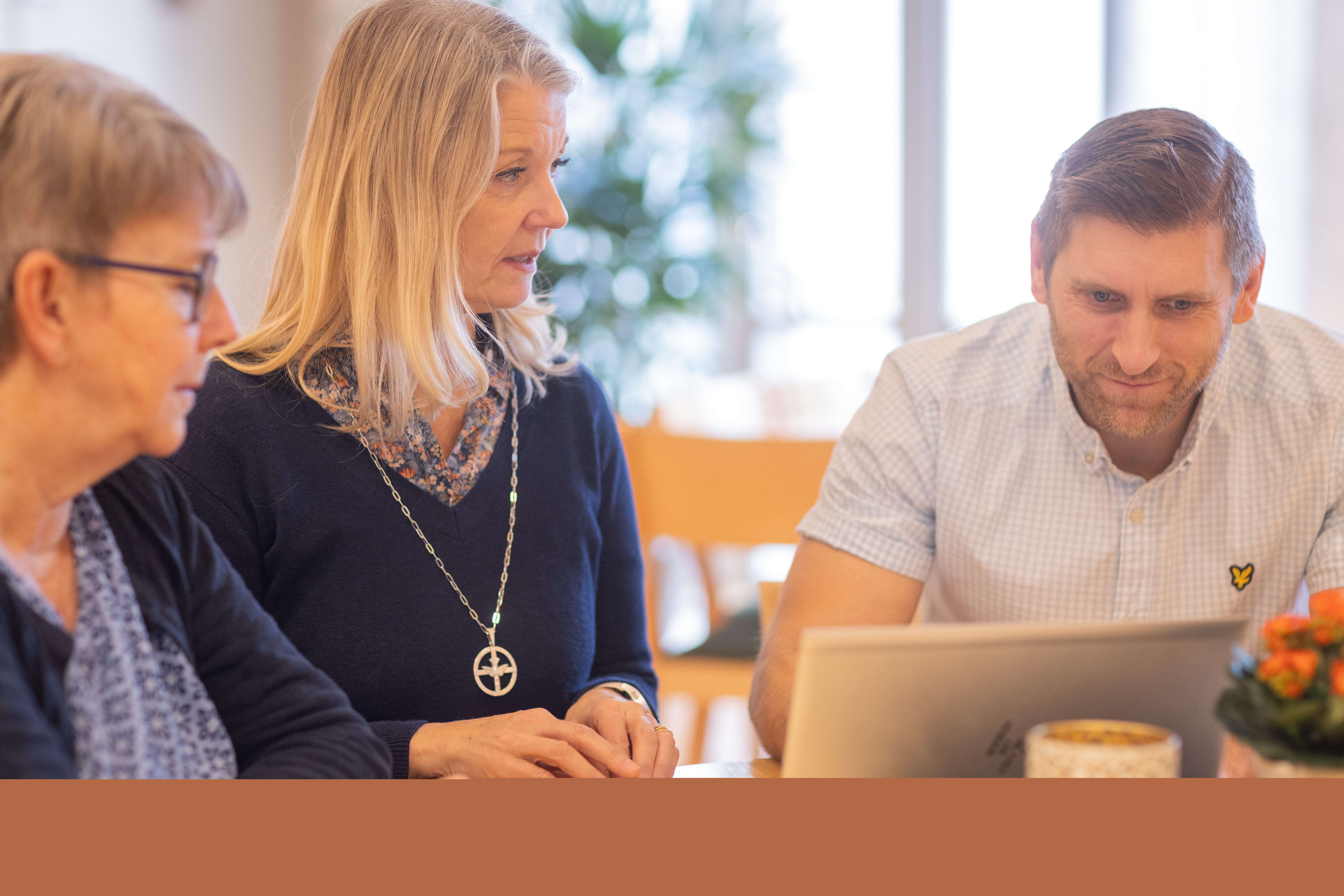 Två kvinnor och en man sitter vid ett bord framför en dator. Oranga blommor och ett värmeljus i förgrunden.