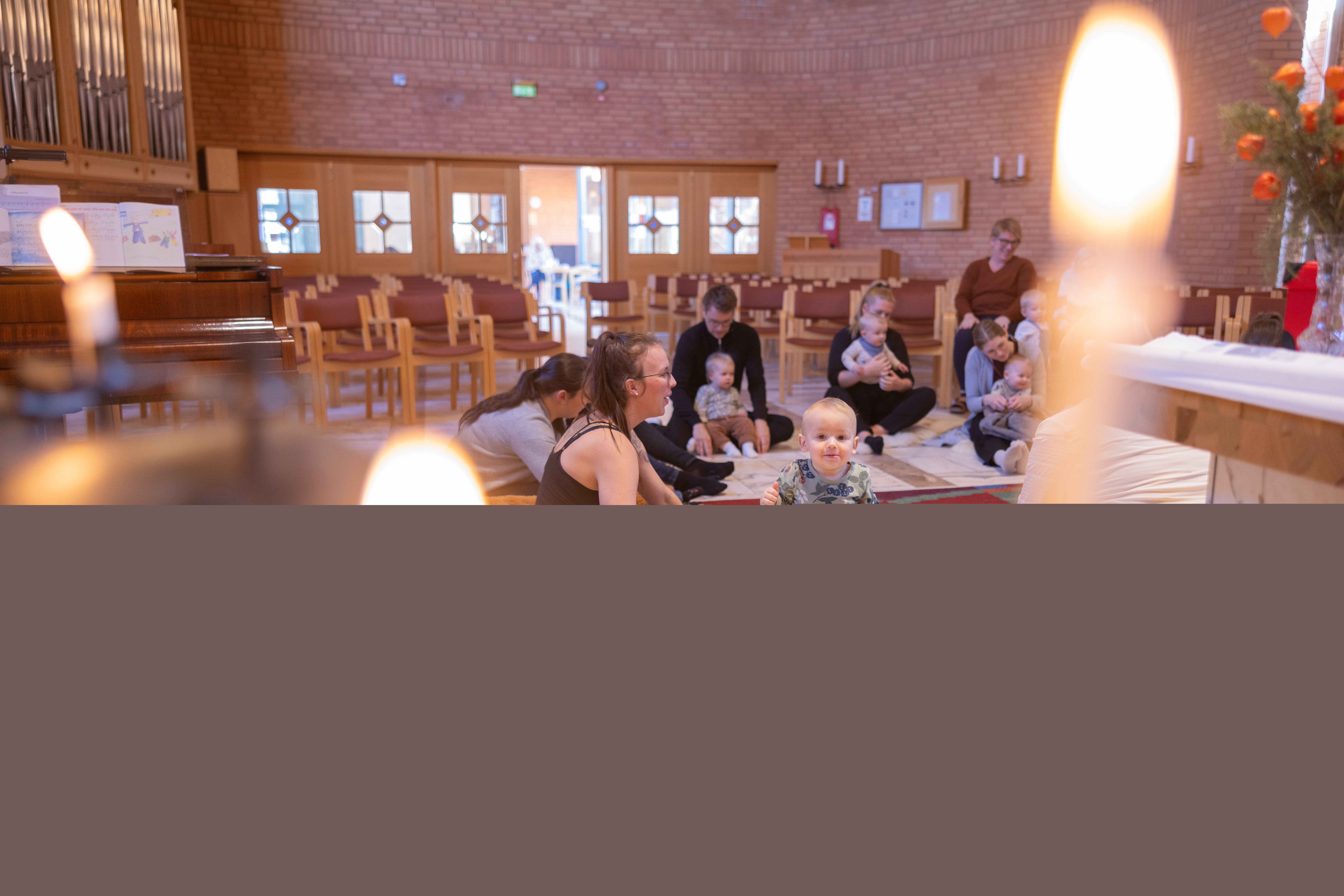 Vuxna och barn sitter på golvet i en kyrka och har sångstund på en öppen förskola. Två tända ljus i förgrunden.