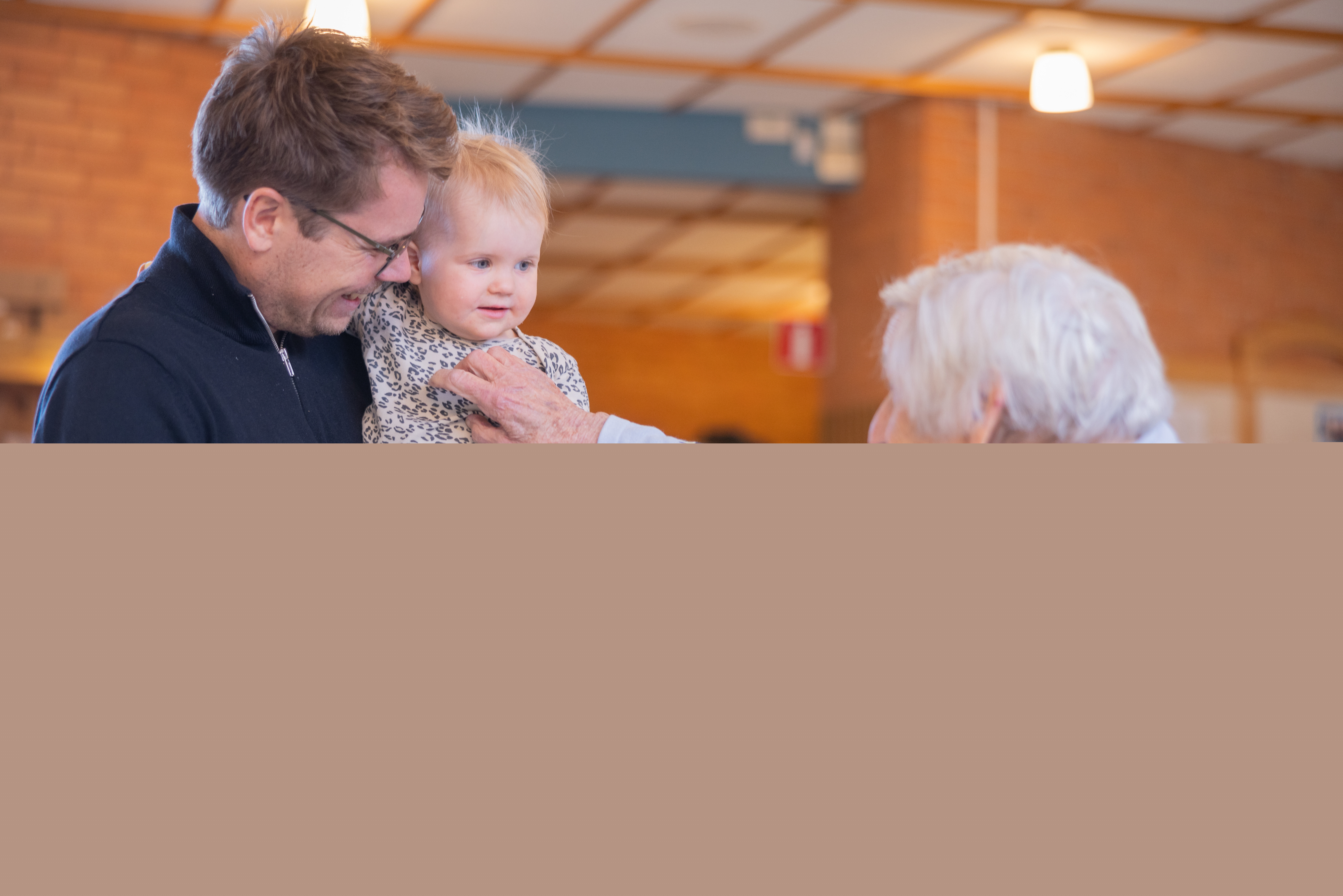 En äldre kvinna busar lekfullt med ett litet barn som sitter i famnen på en vuxen person.