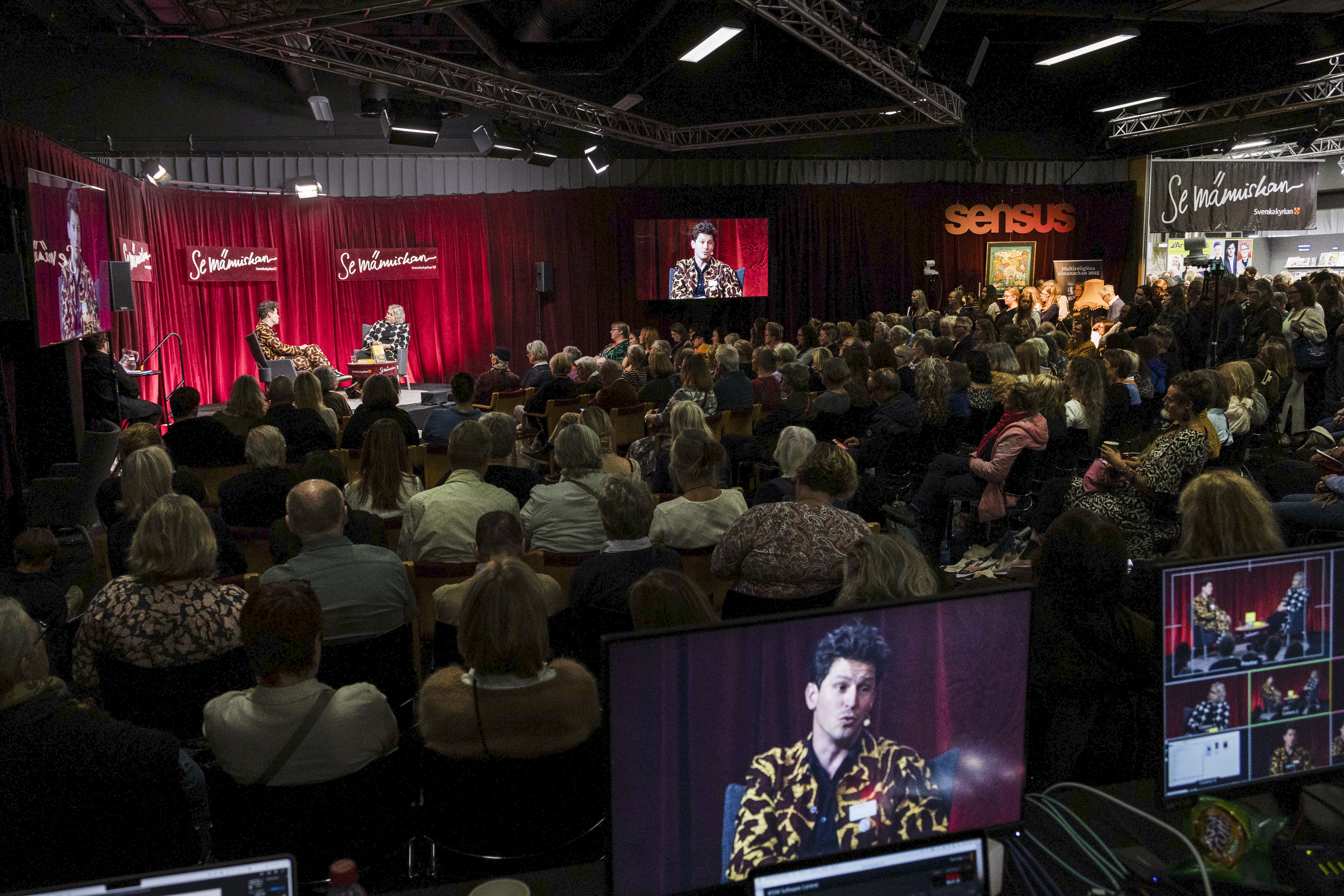 Sittande publik i en lokal på bokmässan. På scenen pågår ett samtal mellan två personer.