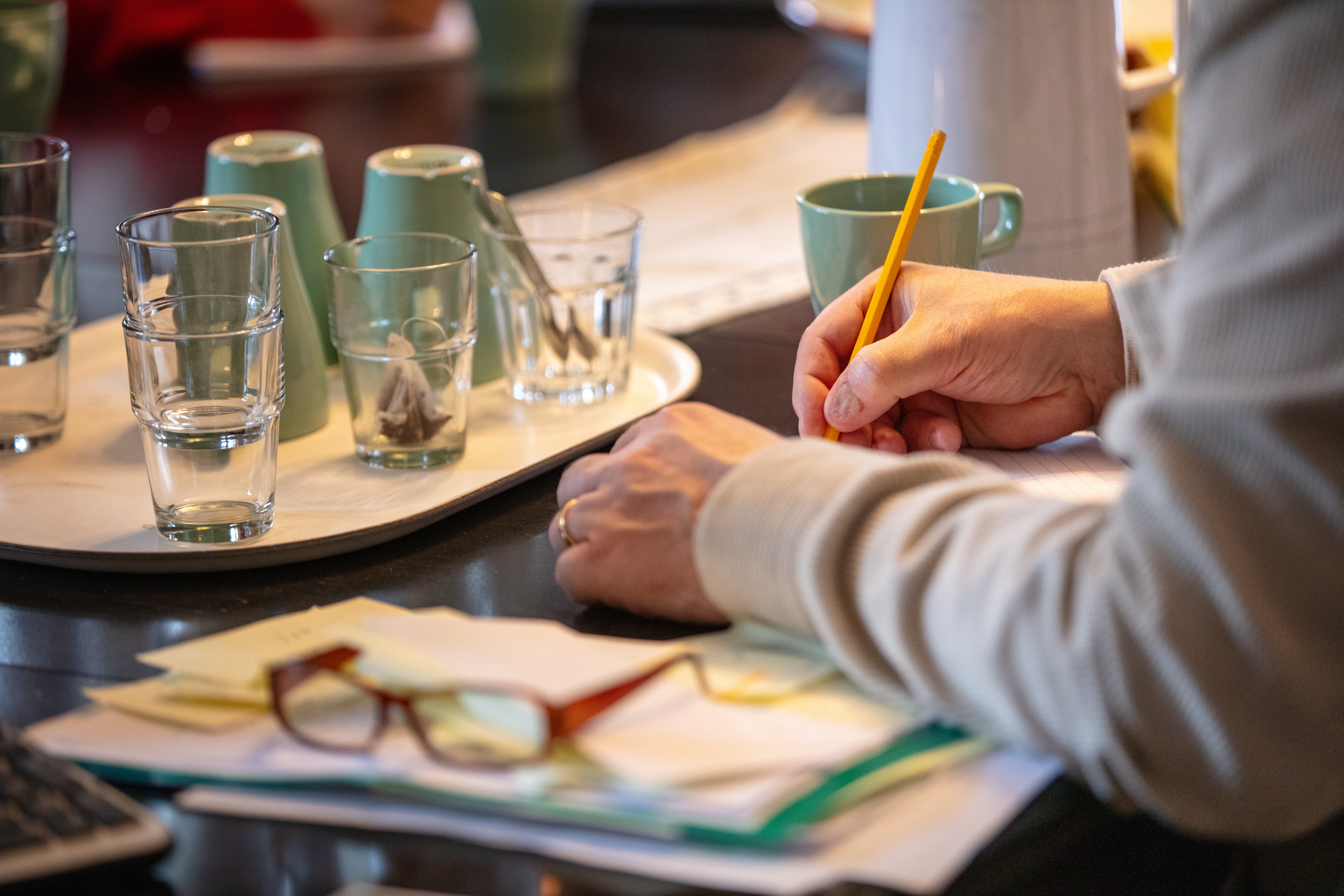 En person håller en penna och skriver något. Personen sitter vid ett bord med en bricka med glas, kaffekoppar och bredvid ligger ett par glasögon.