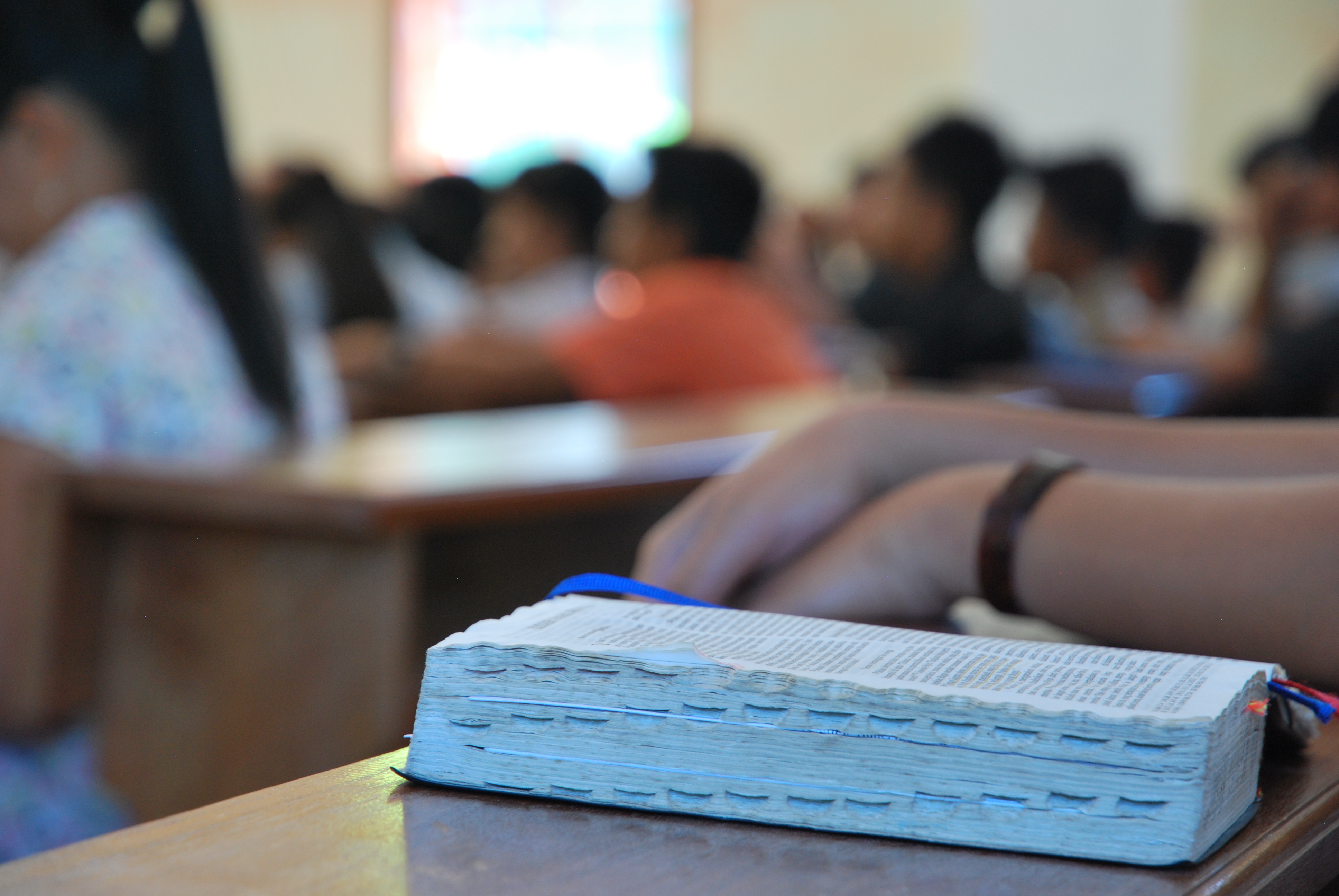 I förgrunden syns en bok ligger på ett bord i ett rum fyllt av människor som sitter i en skolsal. 