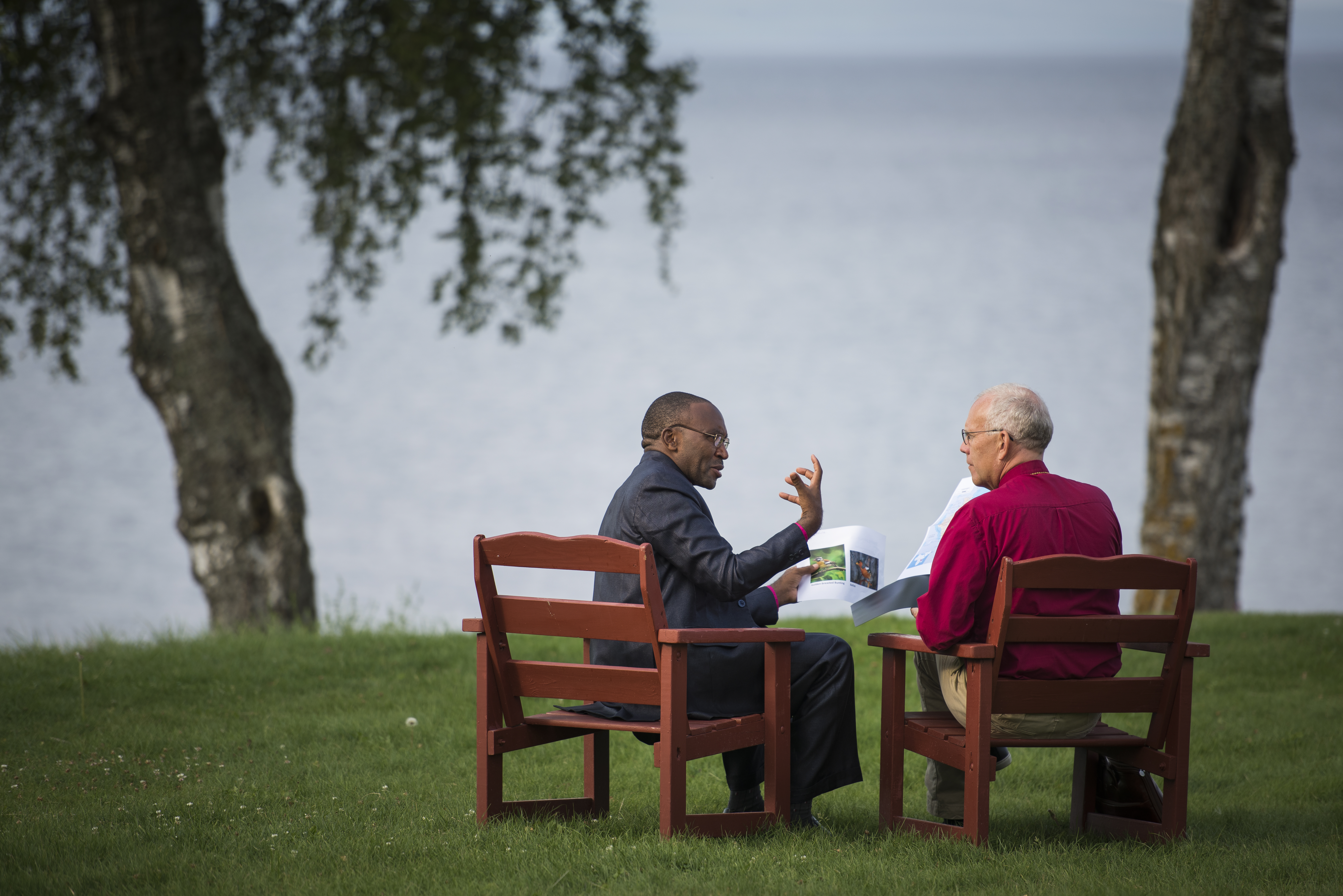 Två personer sitter i stolar på en gräsmatta vid en sjö och pratar med varandra.