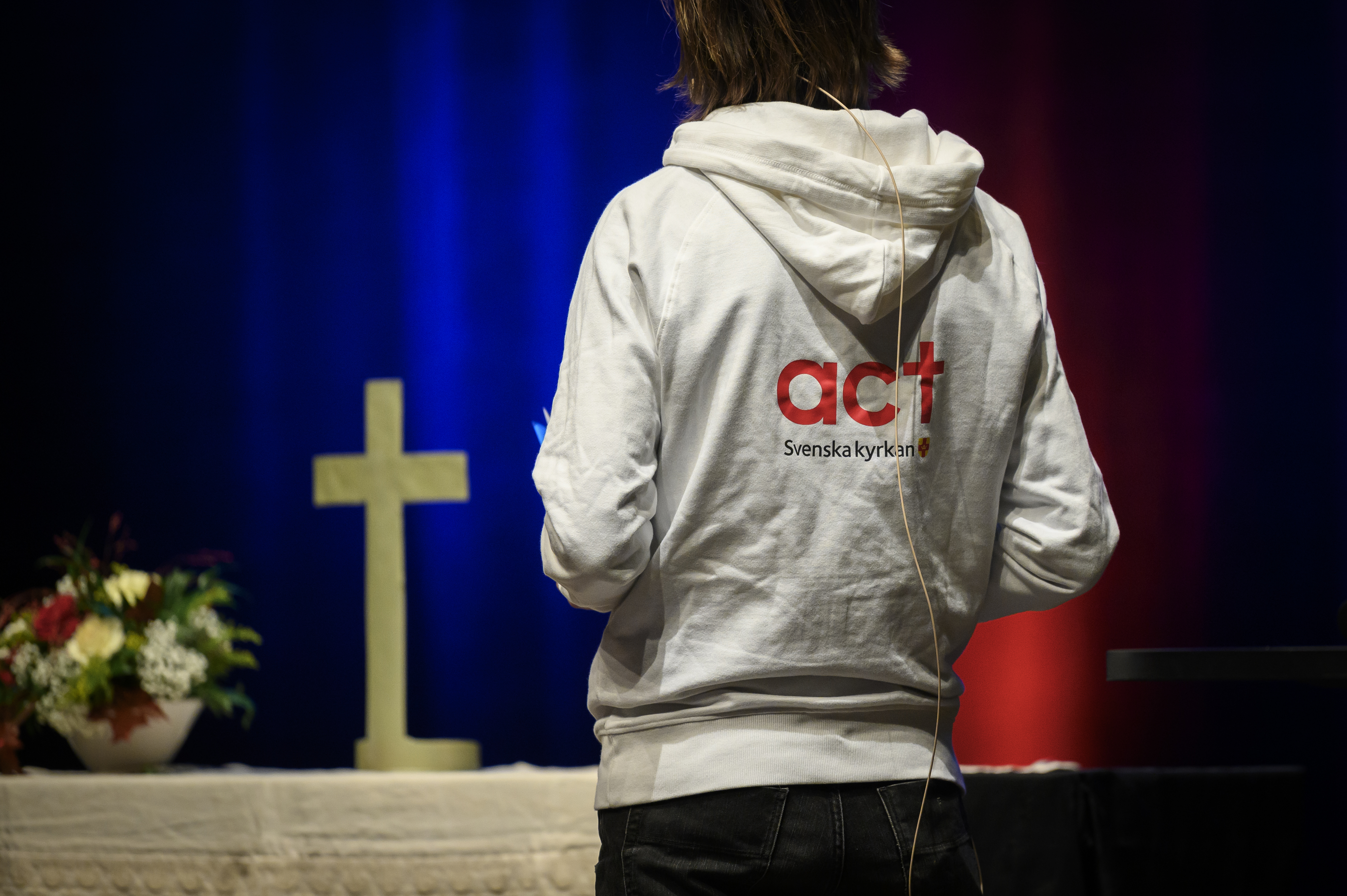Altare med ett kors. Tyggtavlan av en person i luvtröva med texten Act Svenska kyrkan på ryggen. 