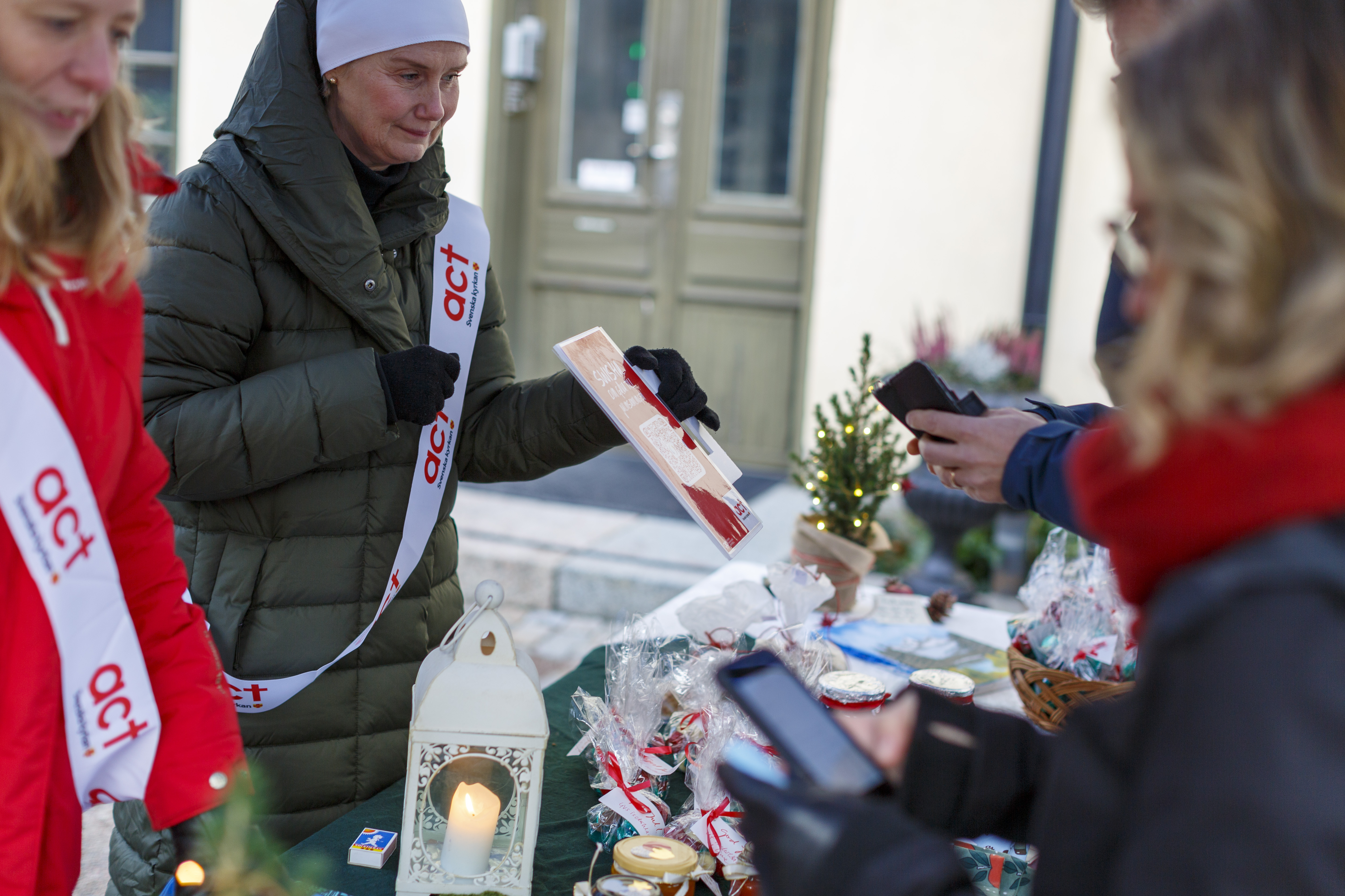 Två kvinnor med Act Svenska kyrkan banderoller över axeln säljer lotter och kakor på julmarknad.