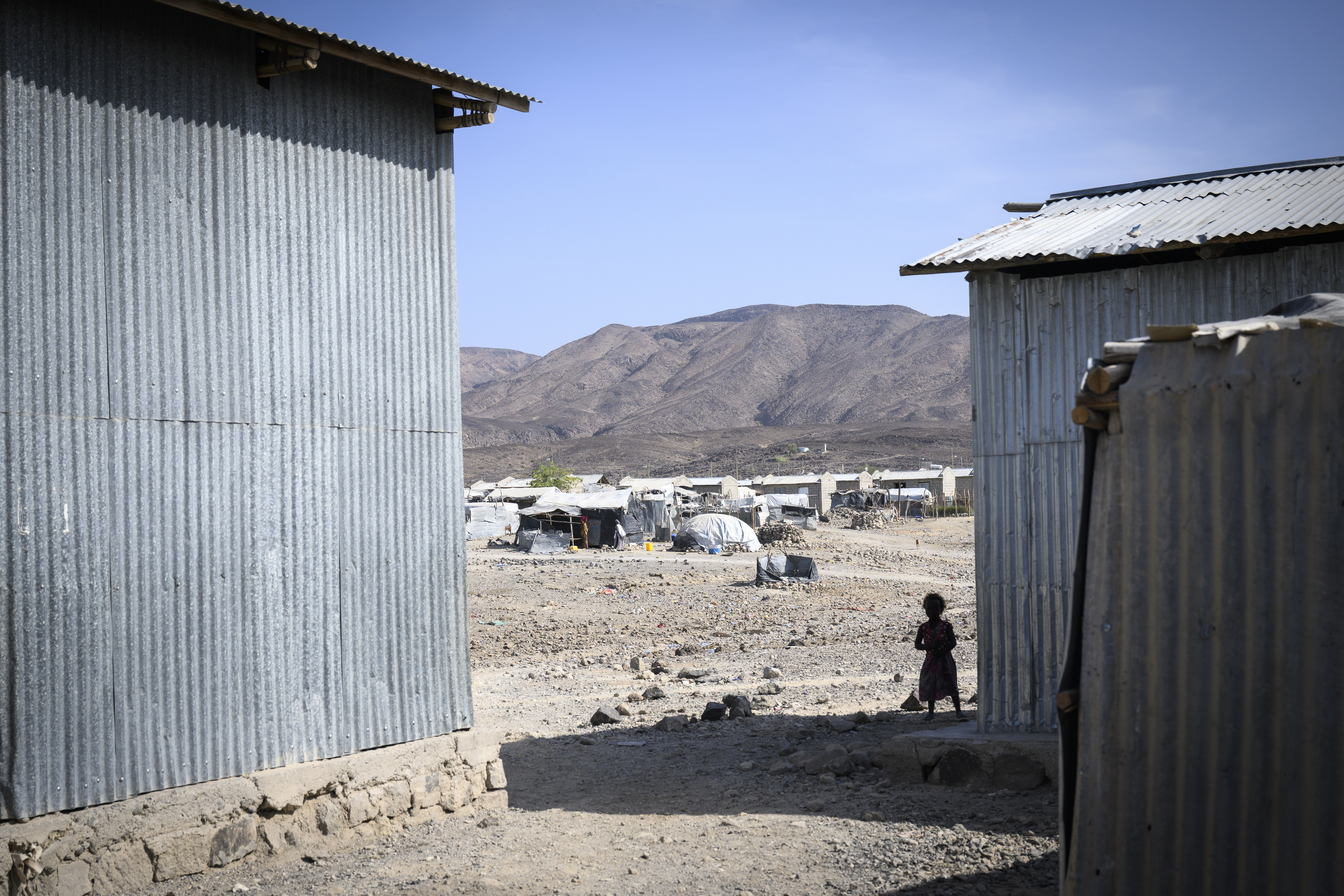 Ett barn mellan två plåtskjul. I bakgrunden syns ett flyktingläger.