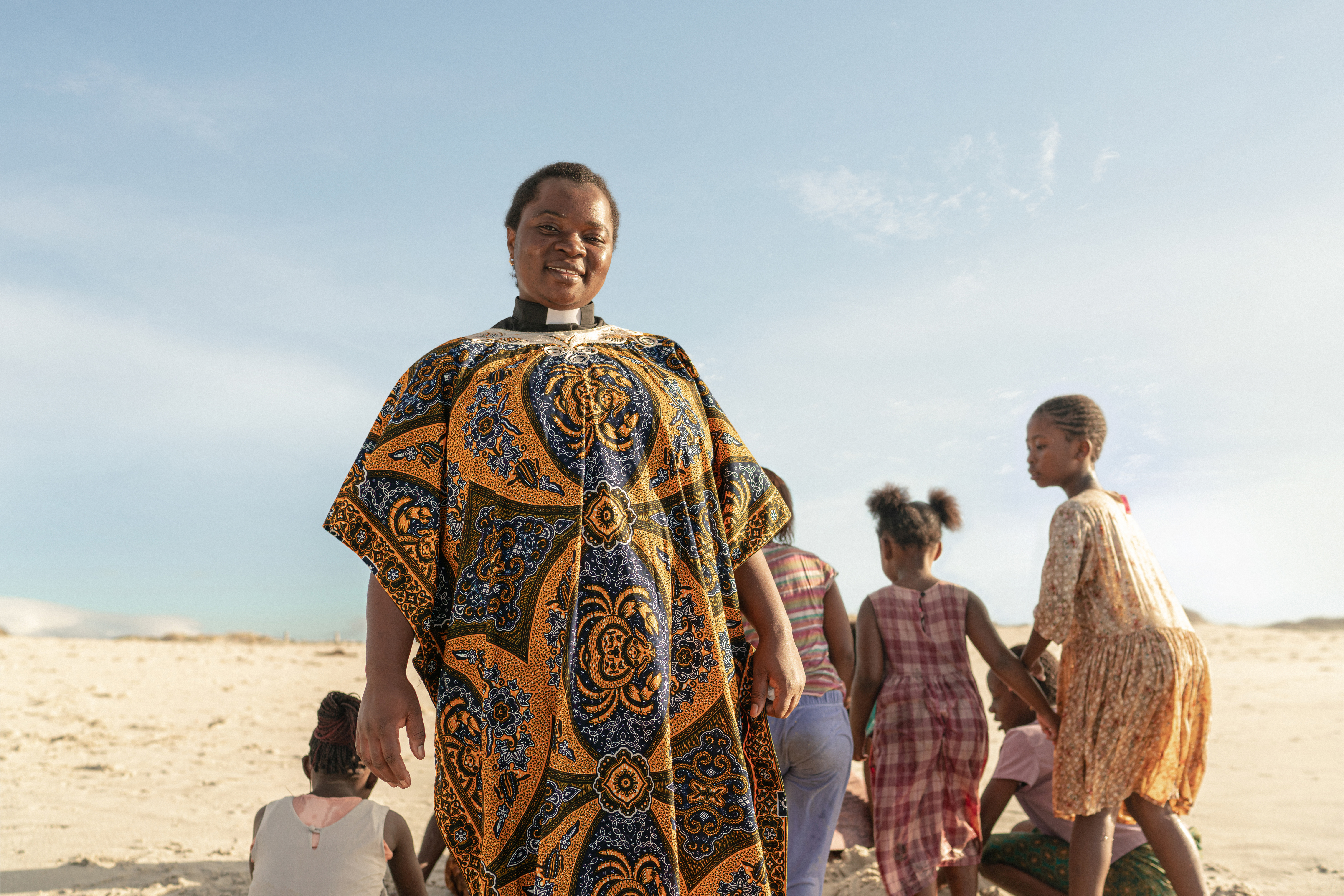 Kvinna i prästkläder på en strand. Bakom henne leker några flickor.