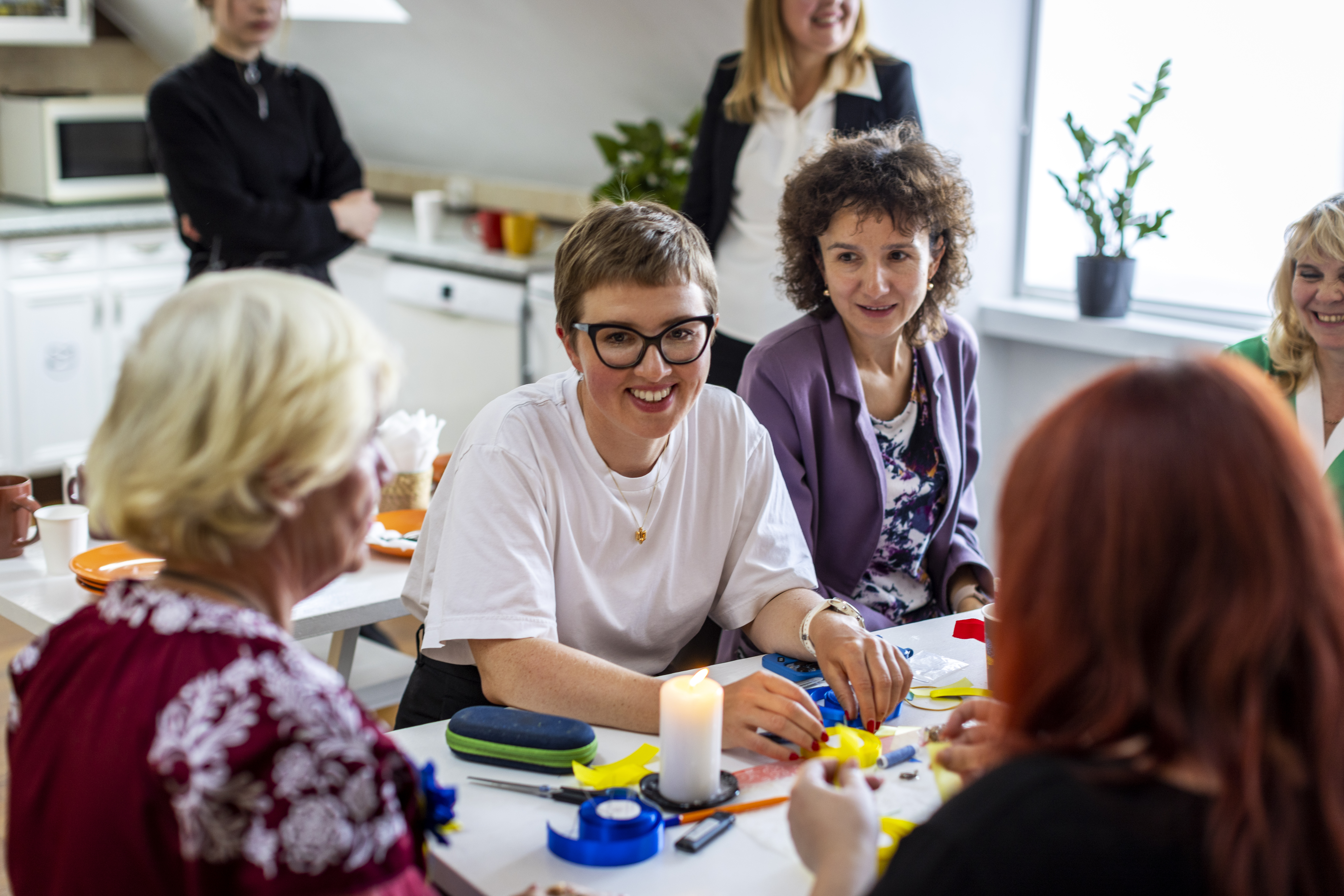 På bilden syns några kvinnor som sitter runt ett bord. De ser glada ut. I bakgrunden syns ett kök. 