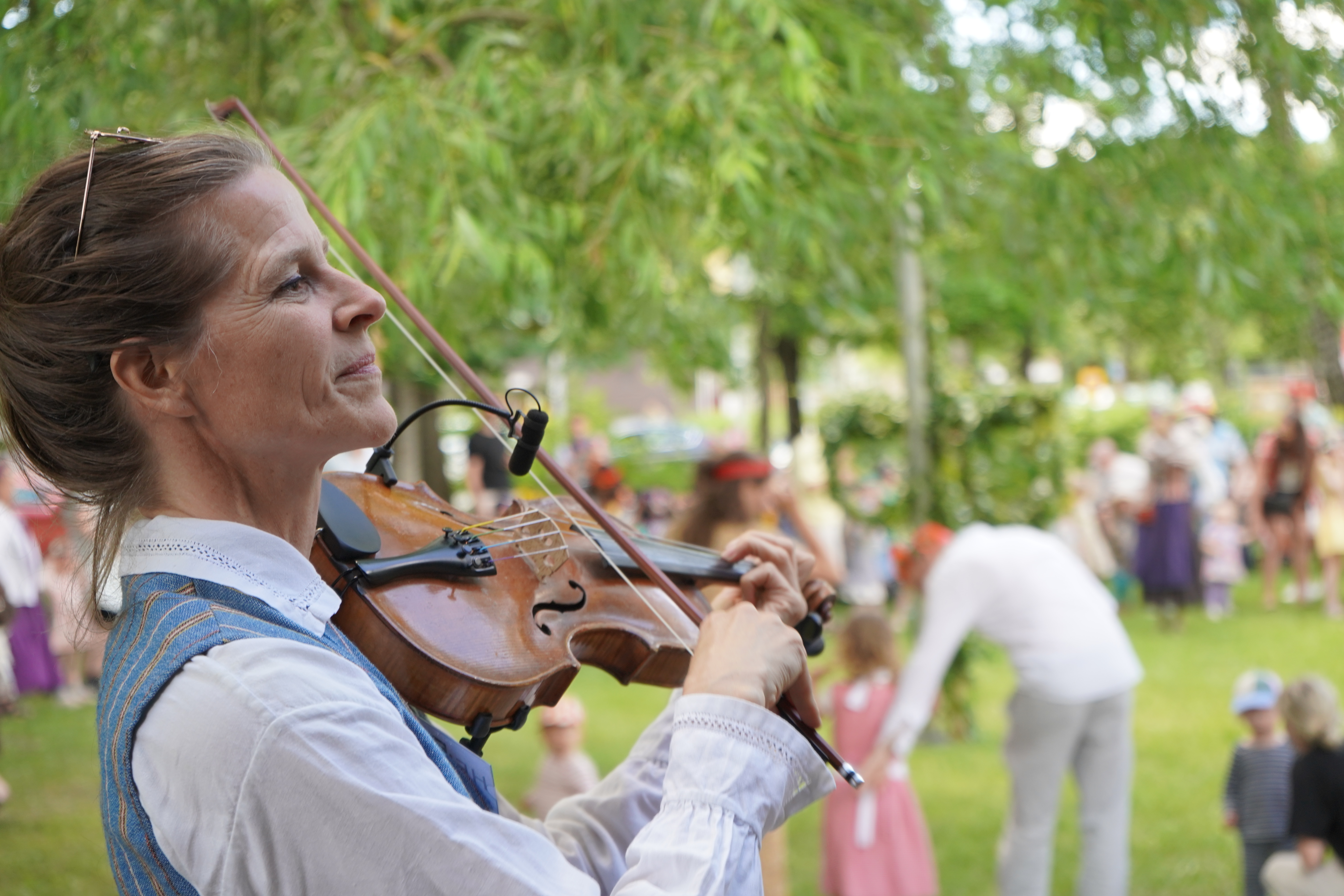En kvinna spelar fiol i folkdräkt. I bakgrunden syns familjer i ett grönområde.