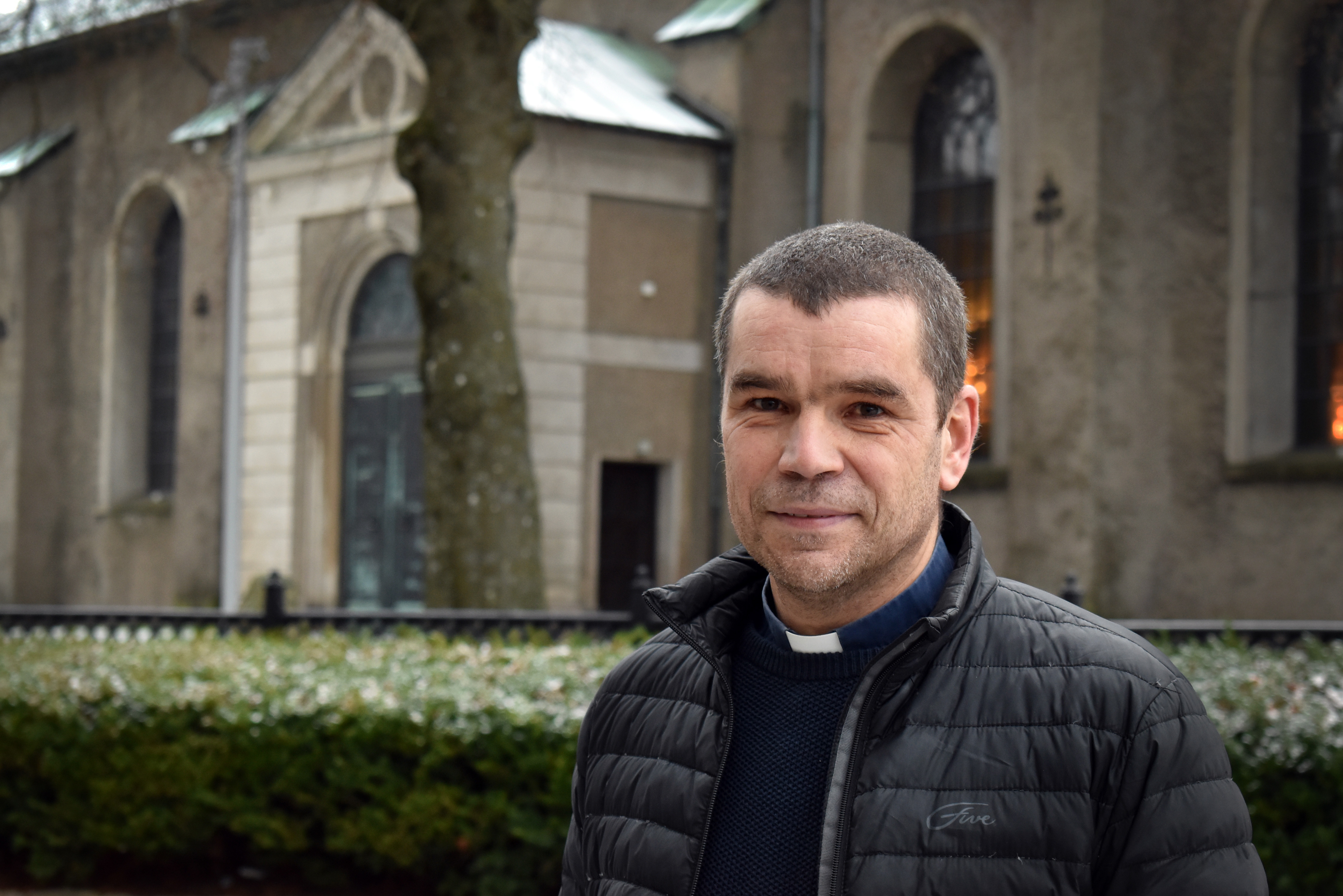 Porträttbild på präst framför kyrkobyggnad. Det är vinter.