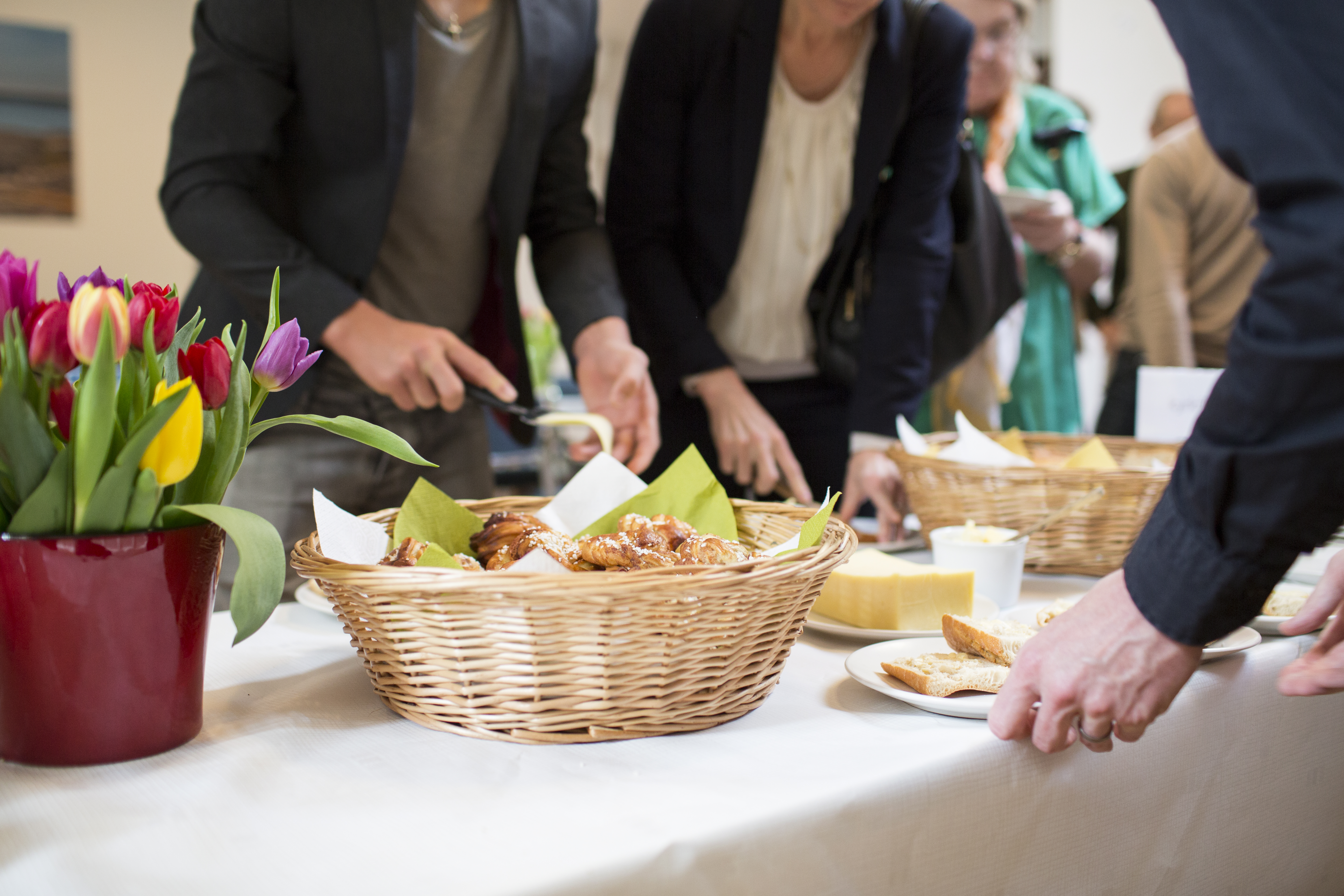 Ett antal personer förser sig från ett bord med bröd, ost och bullar. En färggrann tulpanbukett pryder bordet till vänster