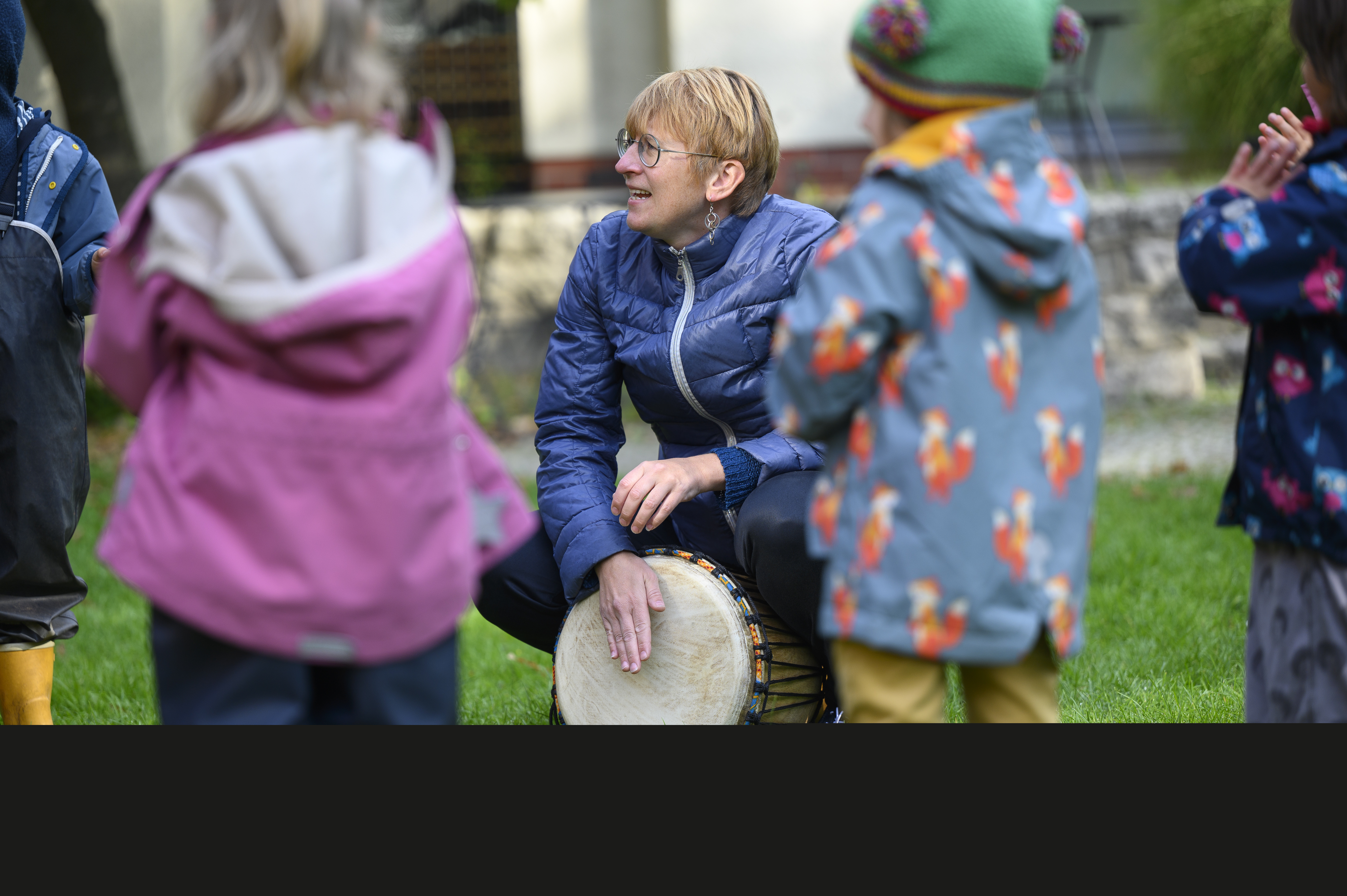 En kvinna sitter på huk utomhus och spelar bongotrumma för en grupp barn.