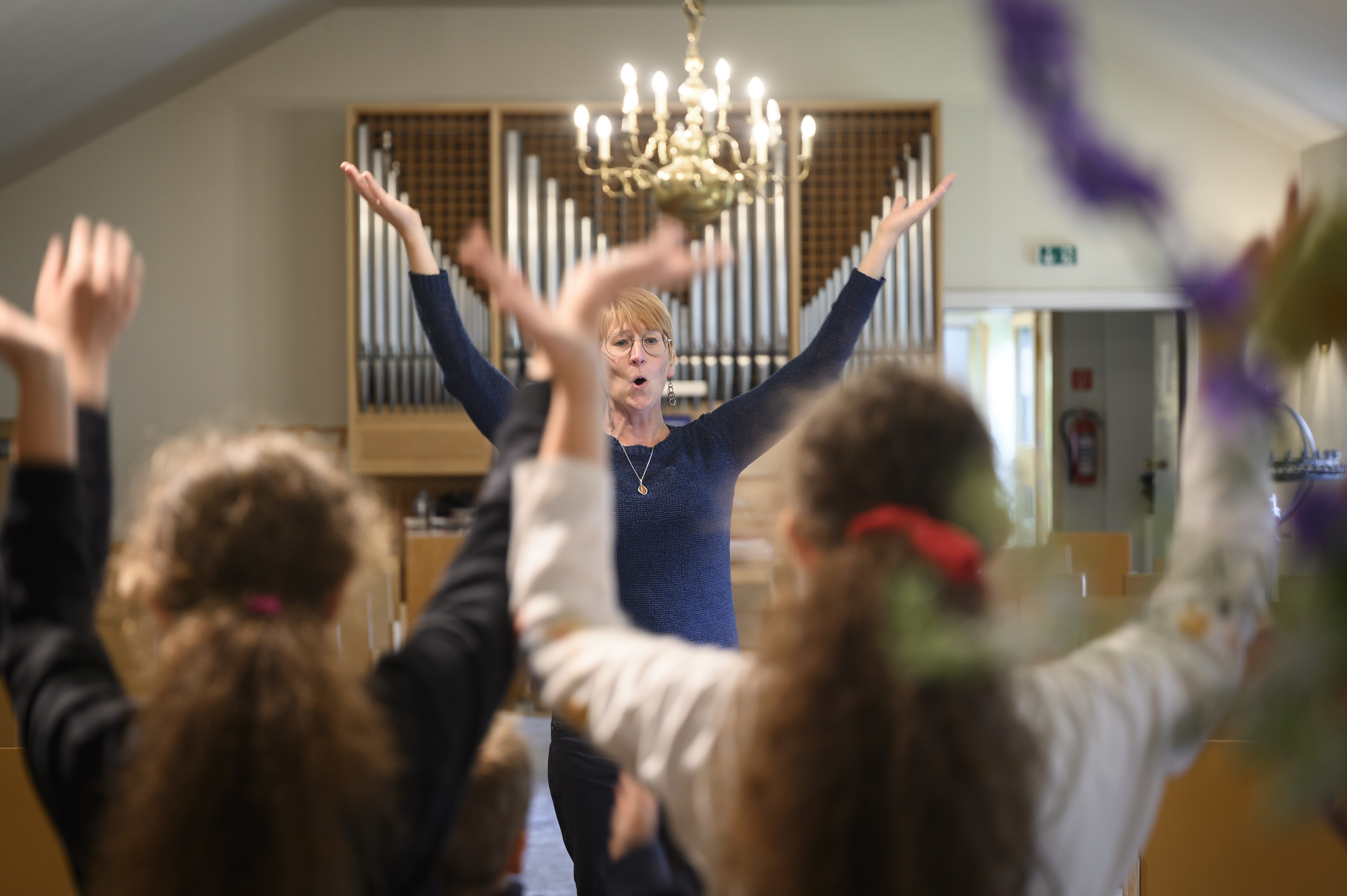 En kvinna står framför en orgel med armarna upp i luften. Några barn står i förgrunden och gör samma gest.