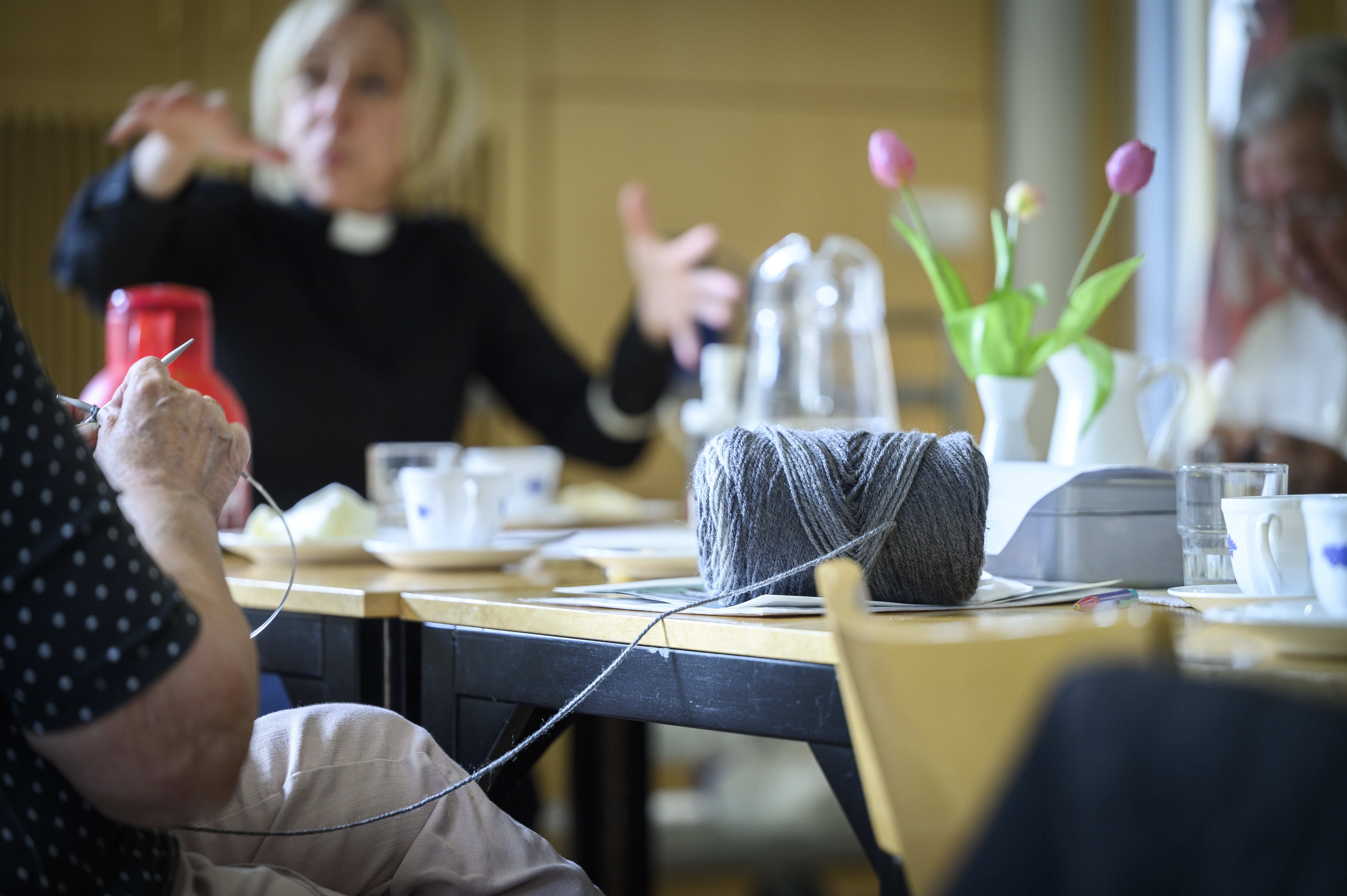 Någon sitter vid ett bord och stickar. Vid bordet syns en kvinnlig präst gestikulera med armarna.